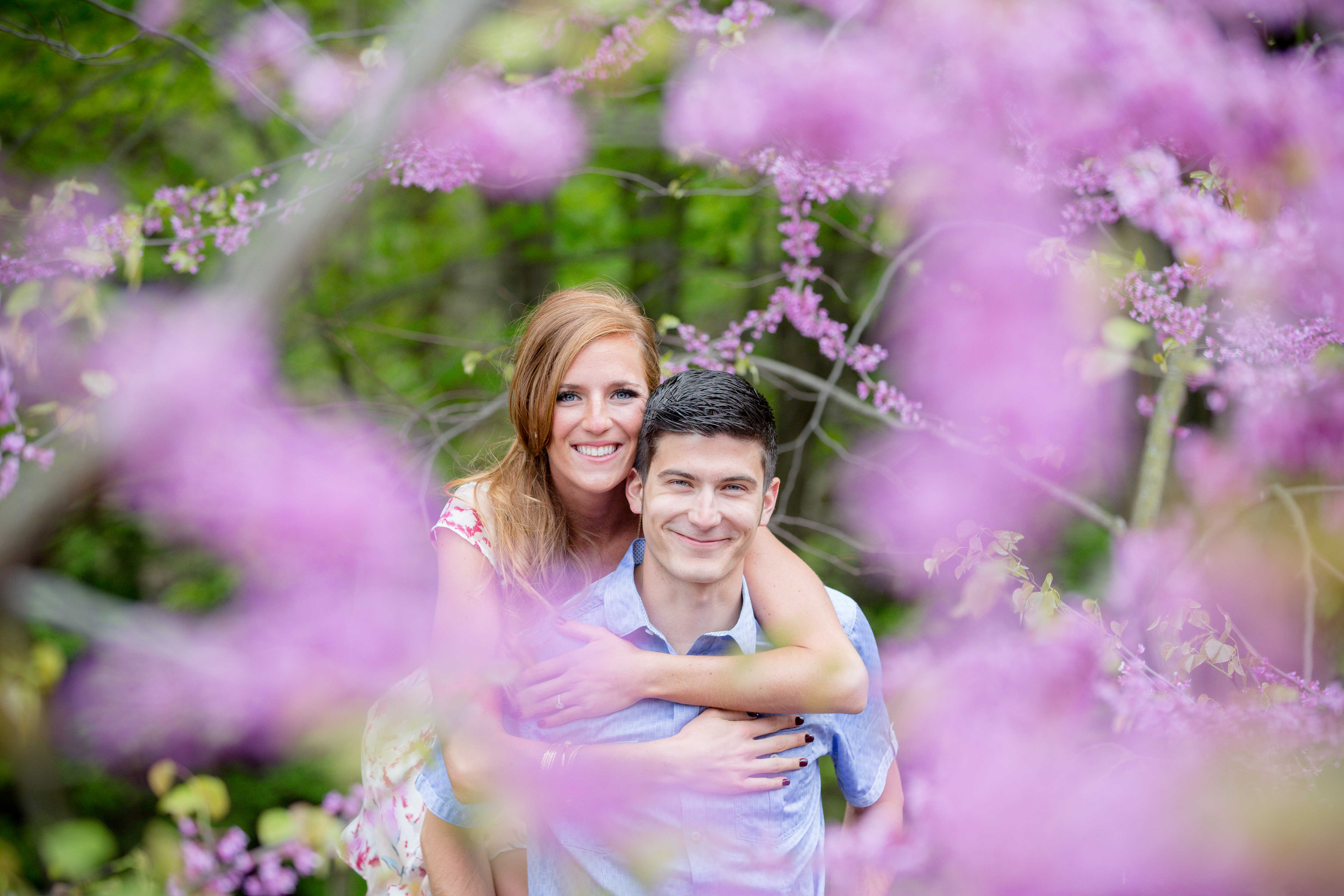 A young woman with red hair and a young man with dark hair smiling, with the woman on the man's back, among pink flowering branches, taken by MN photographer LKW Photography at the MN Landscape Arboretum.