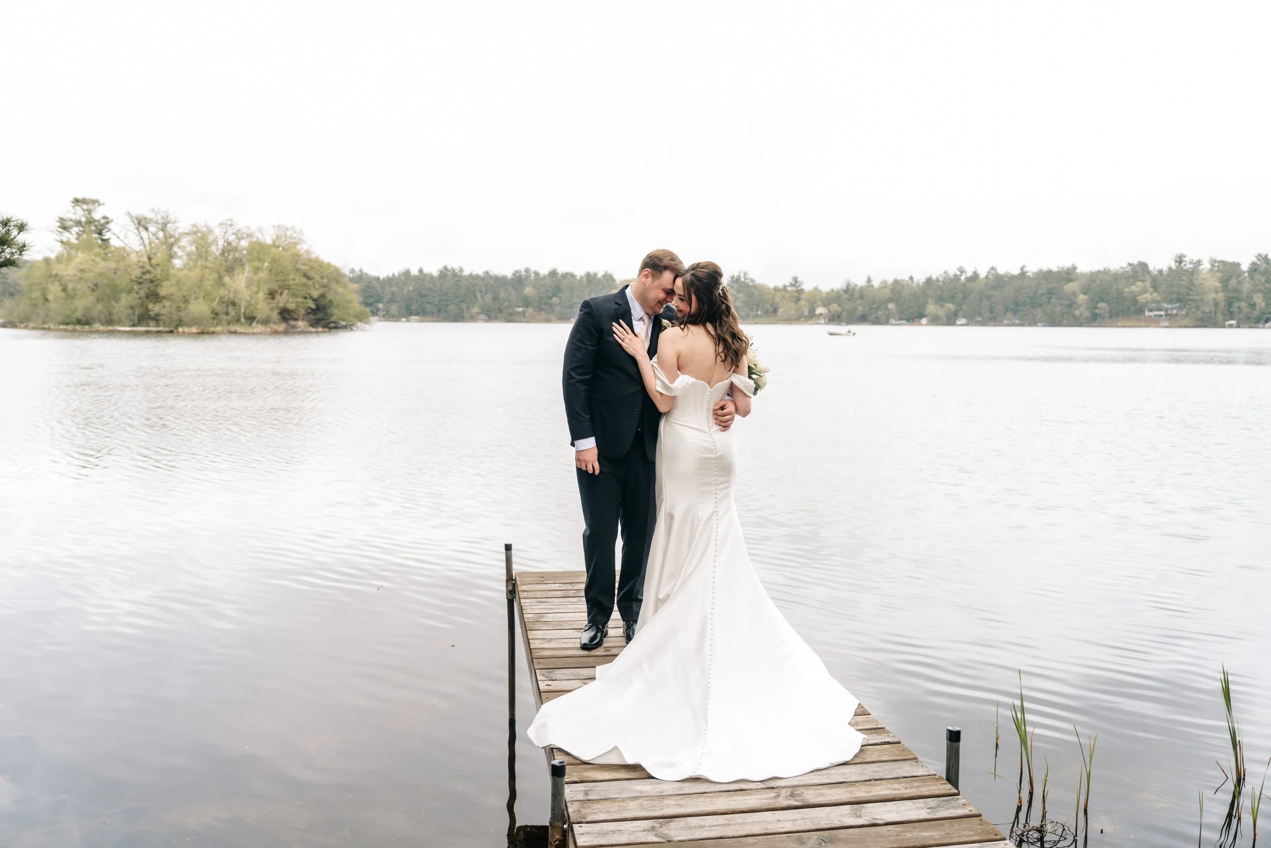 A bride and groom standing close together on a wooden dock by a lake, embracing and smiling. The bride is wearing a white wedding dress, and the groom is in a dark suit. The background features a lake with trees and a cloudy sky.