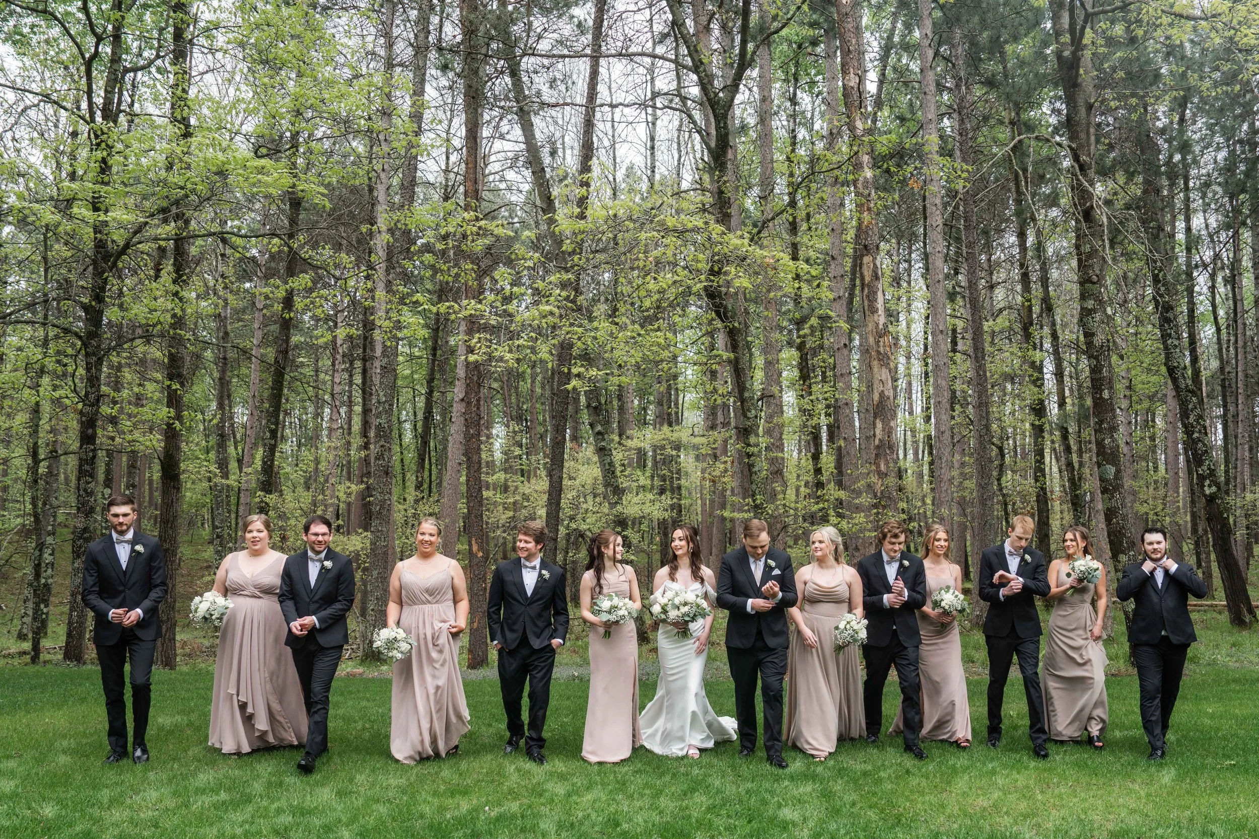 Group of wedding bridesmaids and groomsmen walking across a grassy area in a forest, dressed in formal attire.