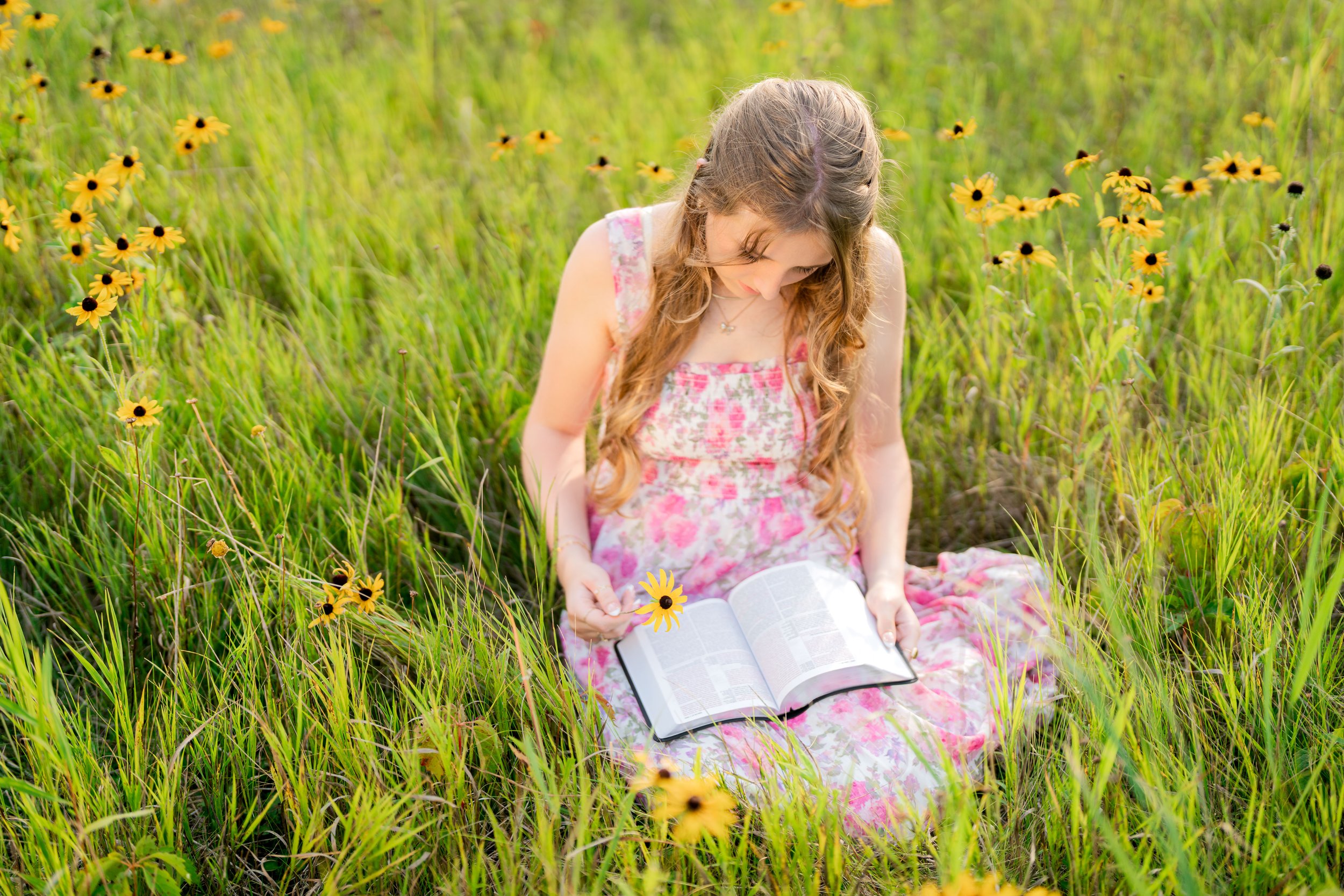 Minnesota senior photographer, LKW Photography, photographs a senior in a pink floral dress sitting in a green meadow with yellow flowers, reading her bible.