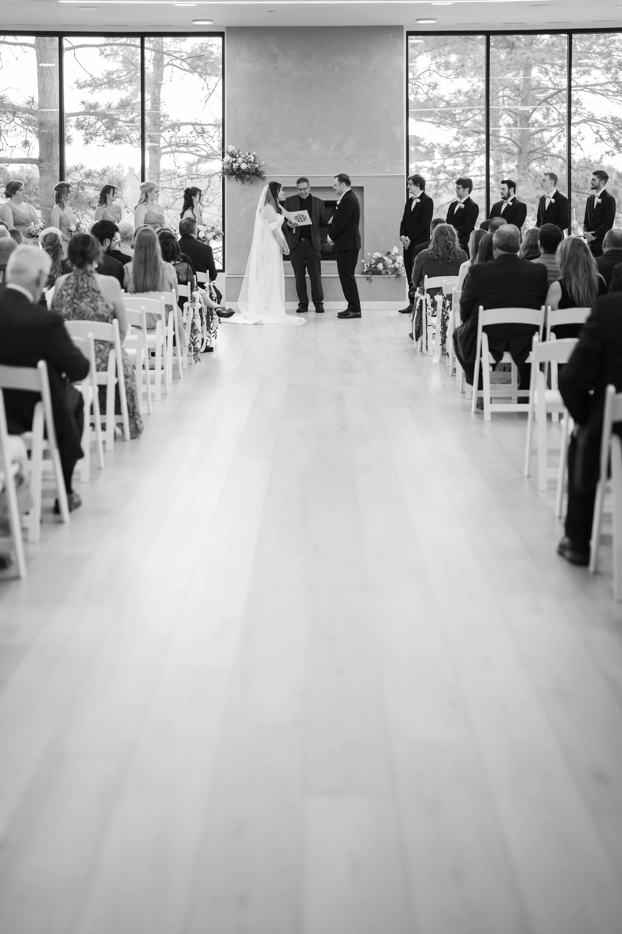 A black-and-white photo of a wedding ceremony inside a bright room with large windows showing trees outside. The bride and groom are standing in front of an officiant, with the wedding party lined up behind them on either side. Guests are seated in w