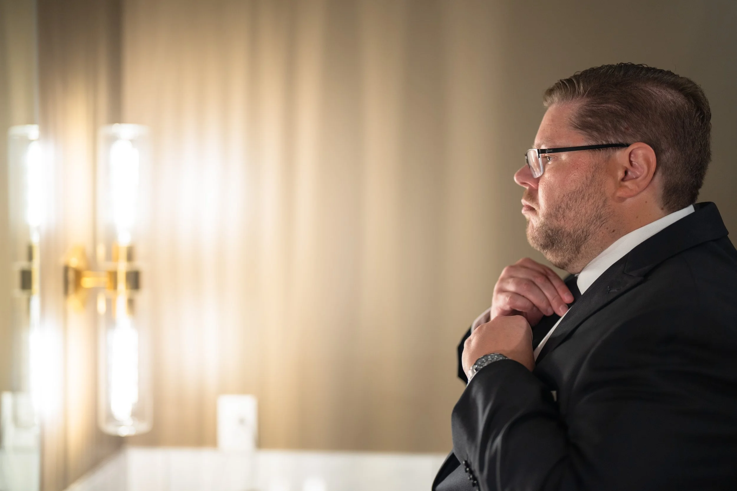 Side profile of a man in a black suit adjusting his tie, with beige curtains and wall-mounted lights in the background, taken by MN photographer LKW Photography.
