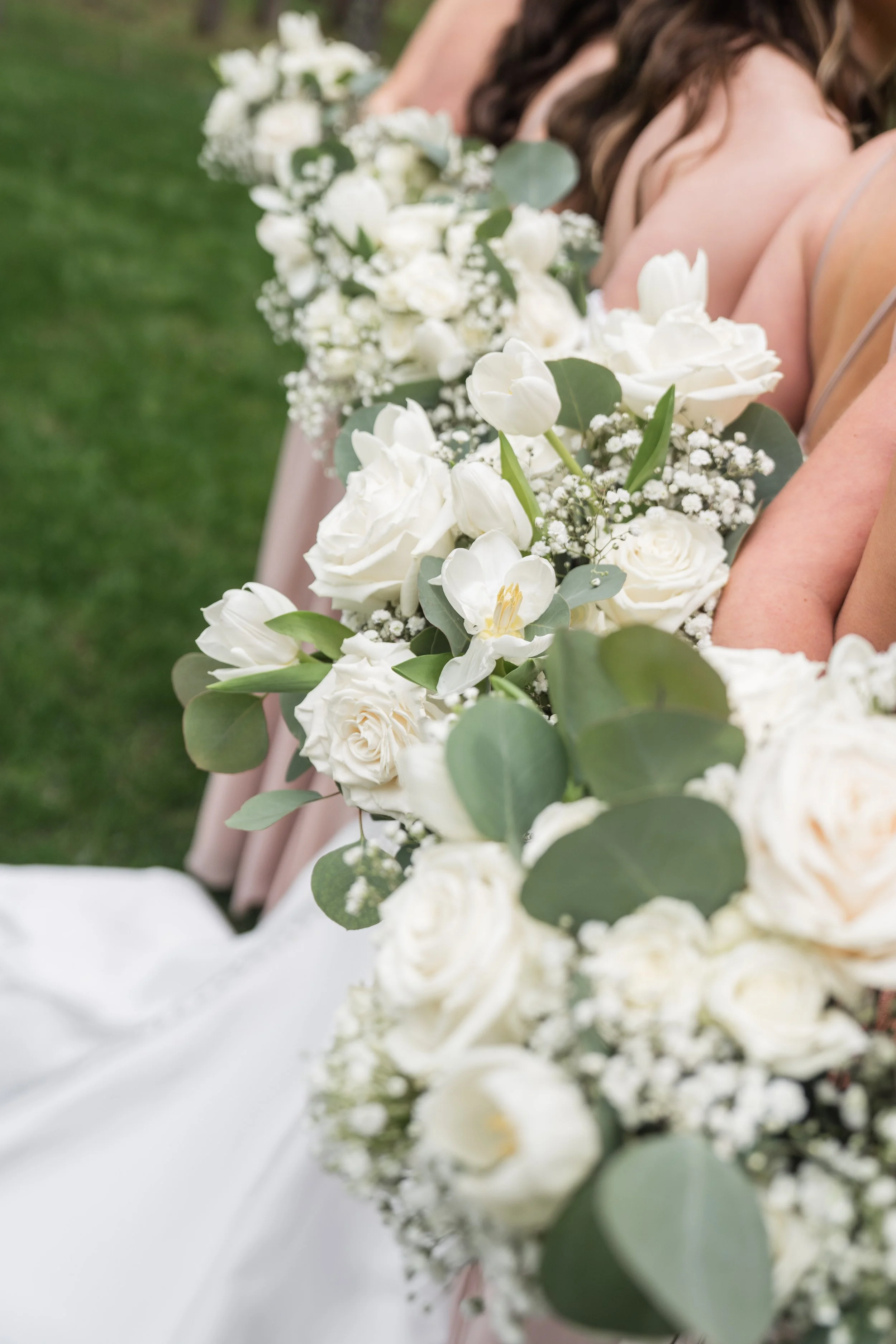 Close-up of a bouquet of white flowers, including roses and tulips, held by women in elegant dresses, likely at a wedding or event, with a grassy background.