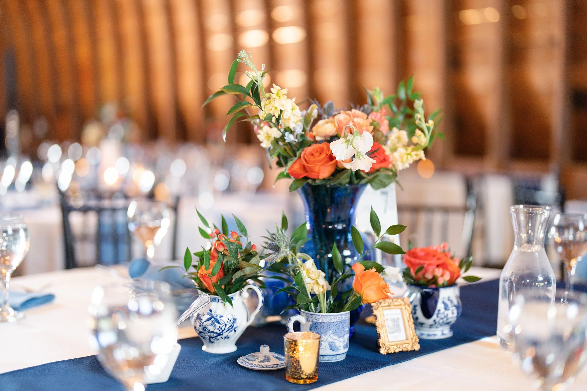 Elegant table centerpiece with a bouquet of white, orange, and pink flowers in a blue and white vase, set for a formal event with plates, glasses, and candles, in a warmly lit venue.