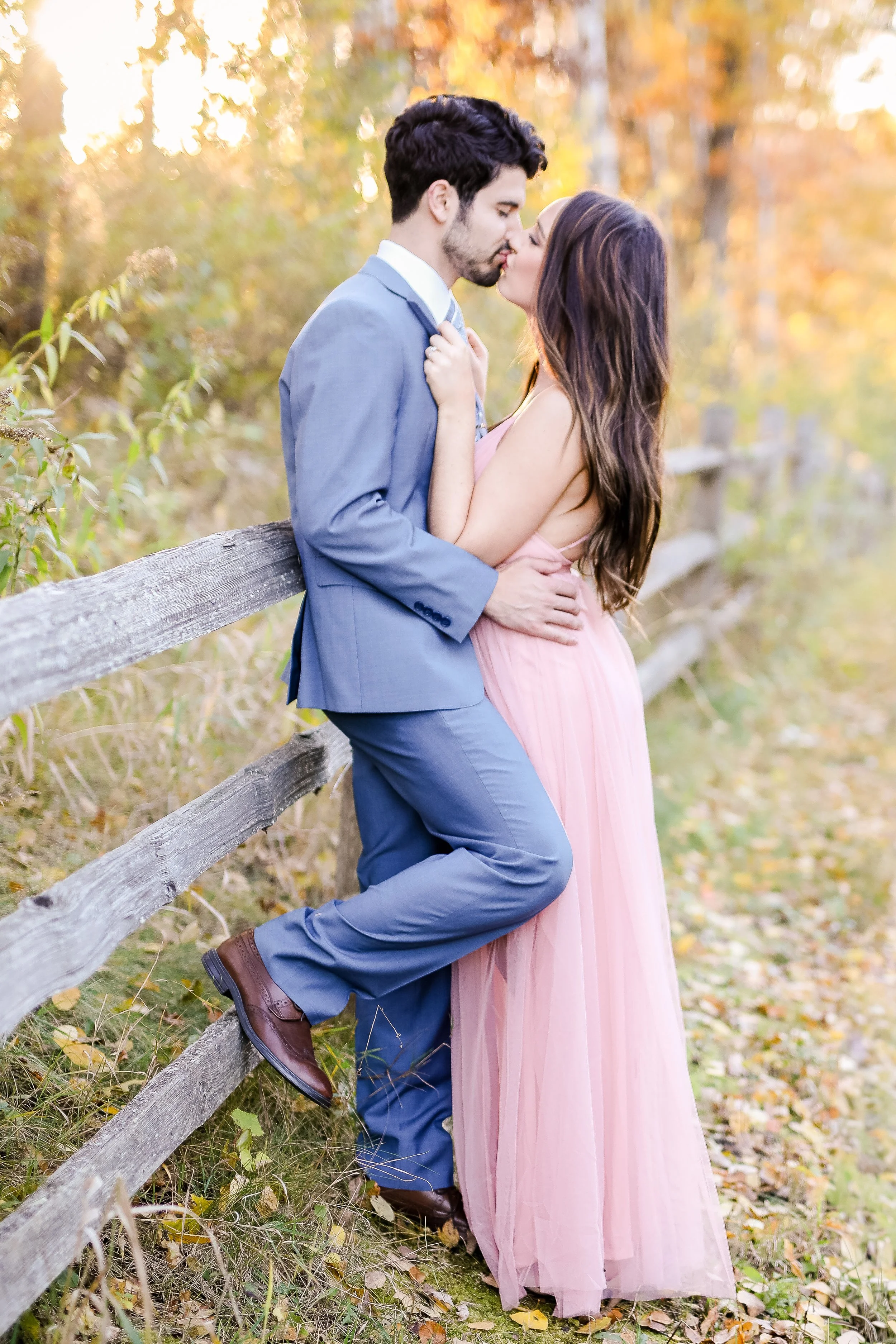 An engaged couple kissing outdoors in autumn, with the man leaning against a wooden fence and the woman standing close to him, dressed in formal attire, taken by Minnesota Wedding Photographer, LKW Photography.