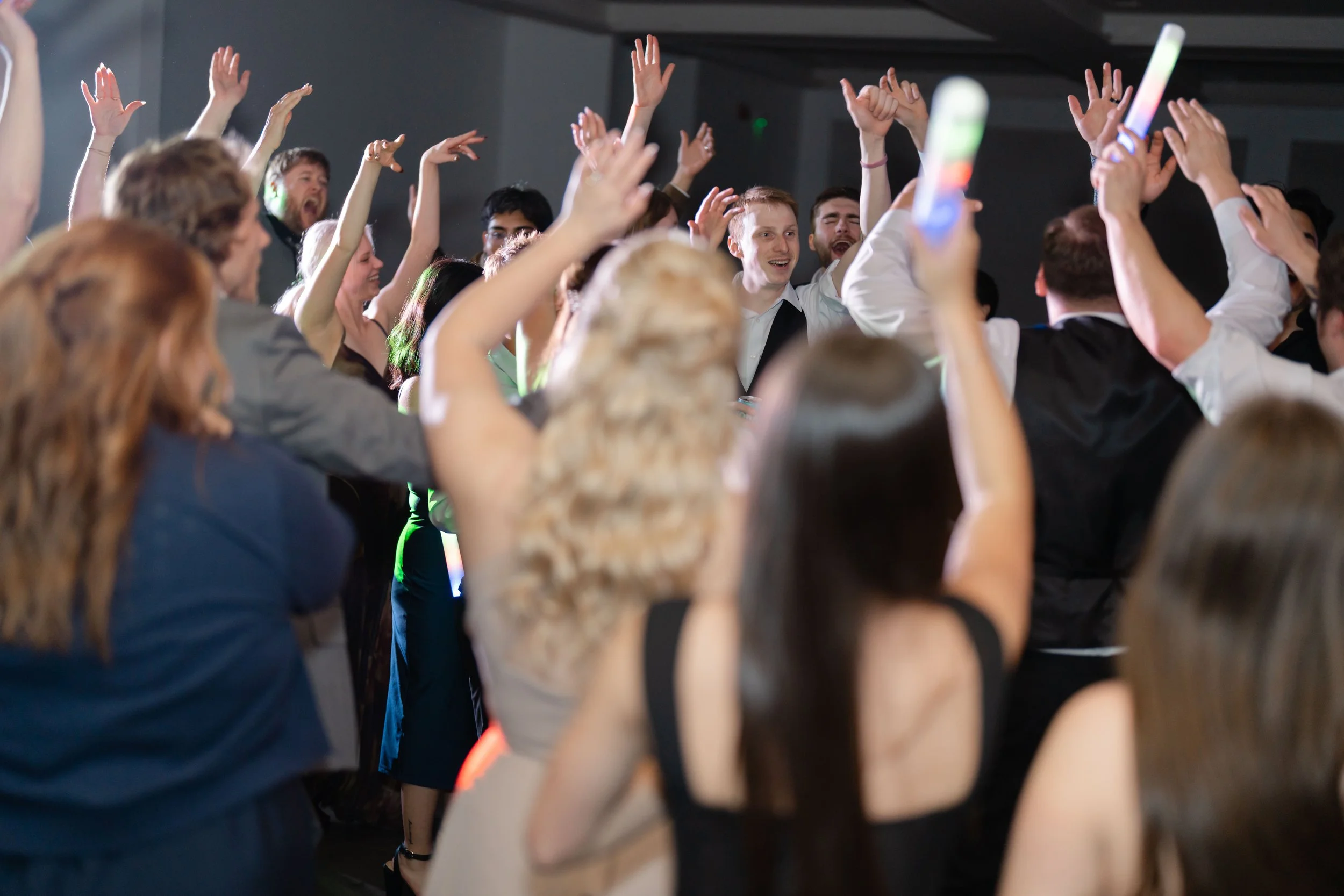 A crowd of people dancing and celebrating at a party or concert, some with their hands raised in the air.