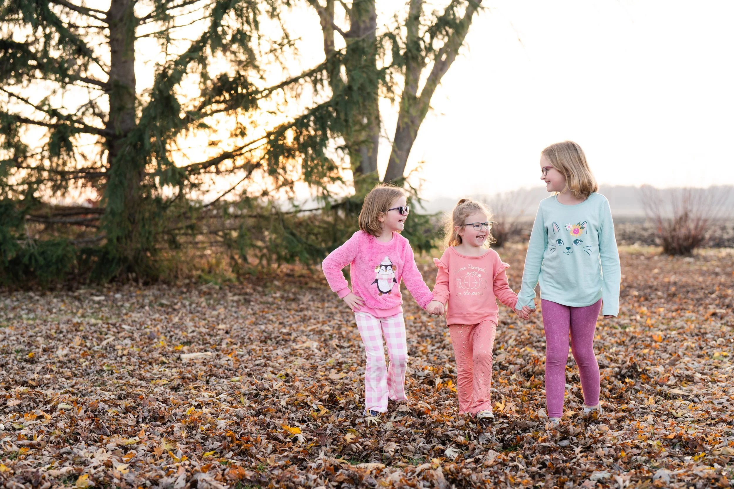 Three young sisters walking hand in hand through a leaf-covered yard during autumn, smiling and enjoying each other's company, taken by MN photographer LKW Photography.