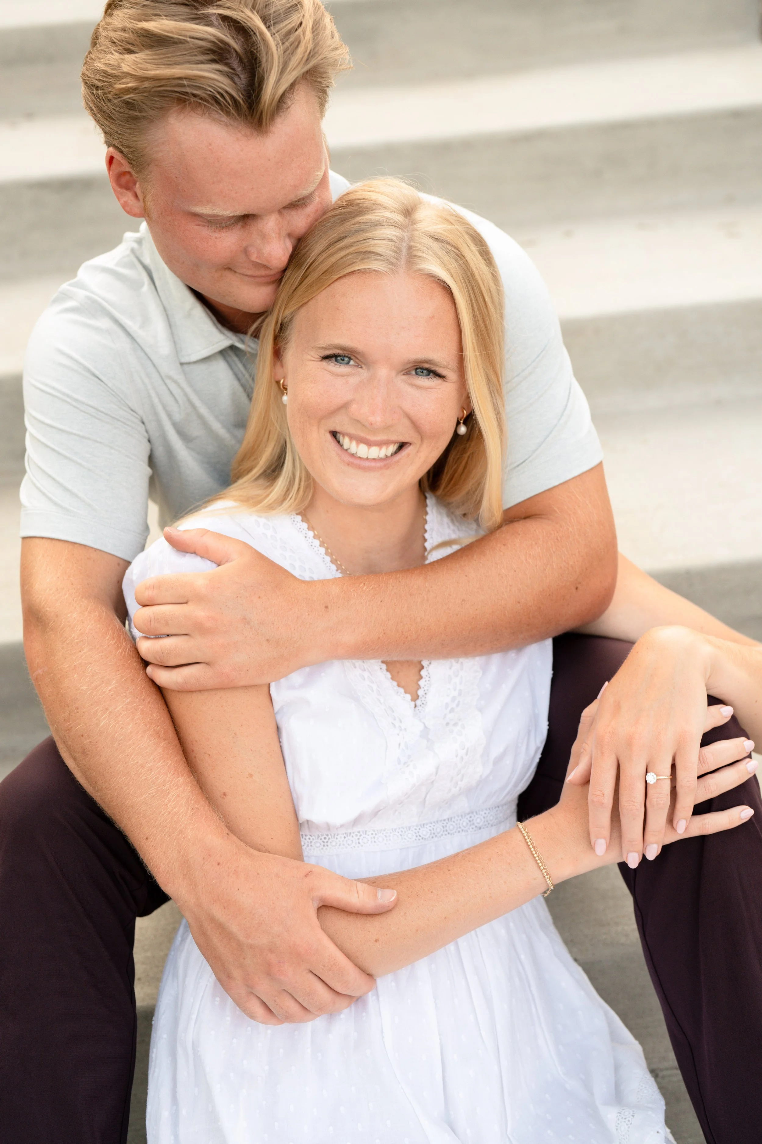 A beautiful engagement photo at The Port of Excelsior in MN.  The couple is sitting on stairs, with the bride to be looking at the camera, and the groom to be sitting behind her, his arms wrapped around her, as he looks down at her.