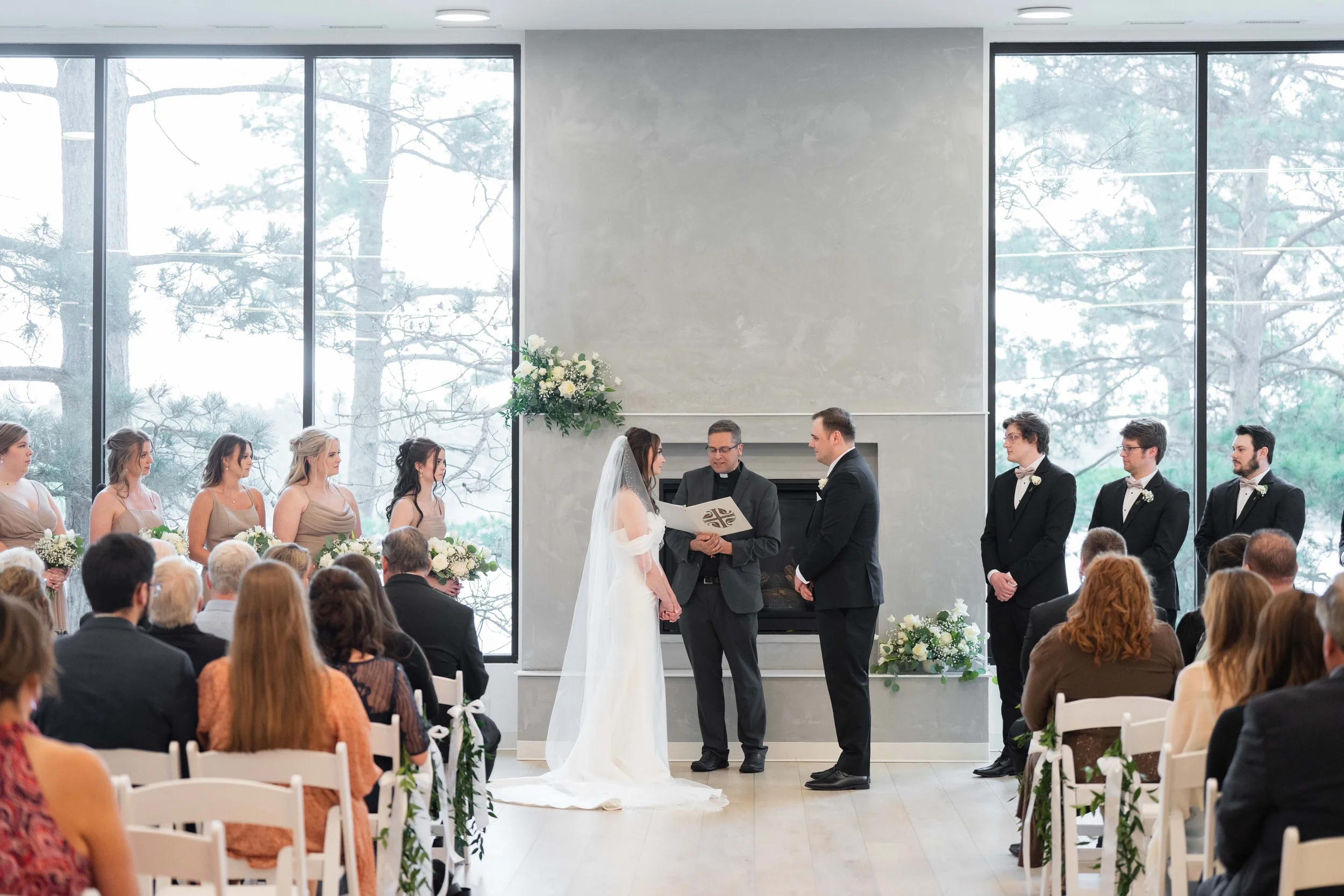 A wedding ceremony taking place indoors with large windows showing trees outside. The bride and groom are standing before an officiant, exchanging vows. Bridesmaids are on the left, and groomsmen are on the right, all dressed formally. Guests are sea