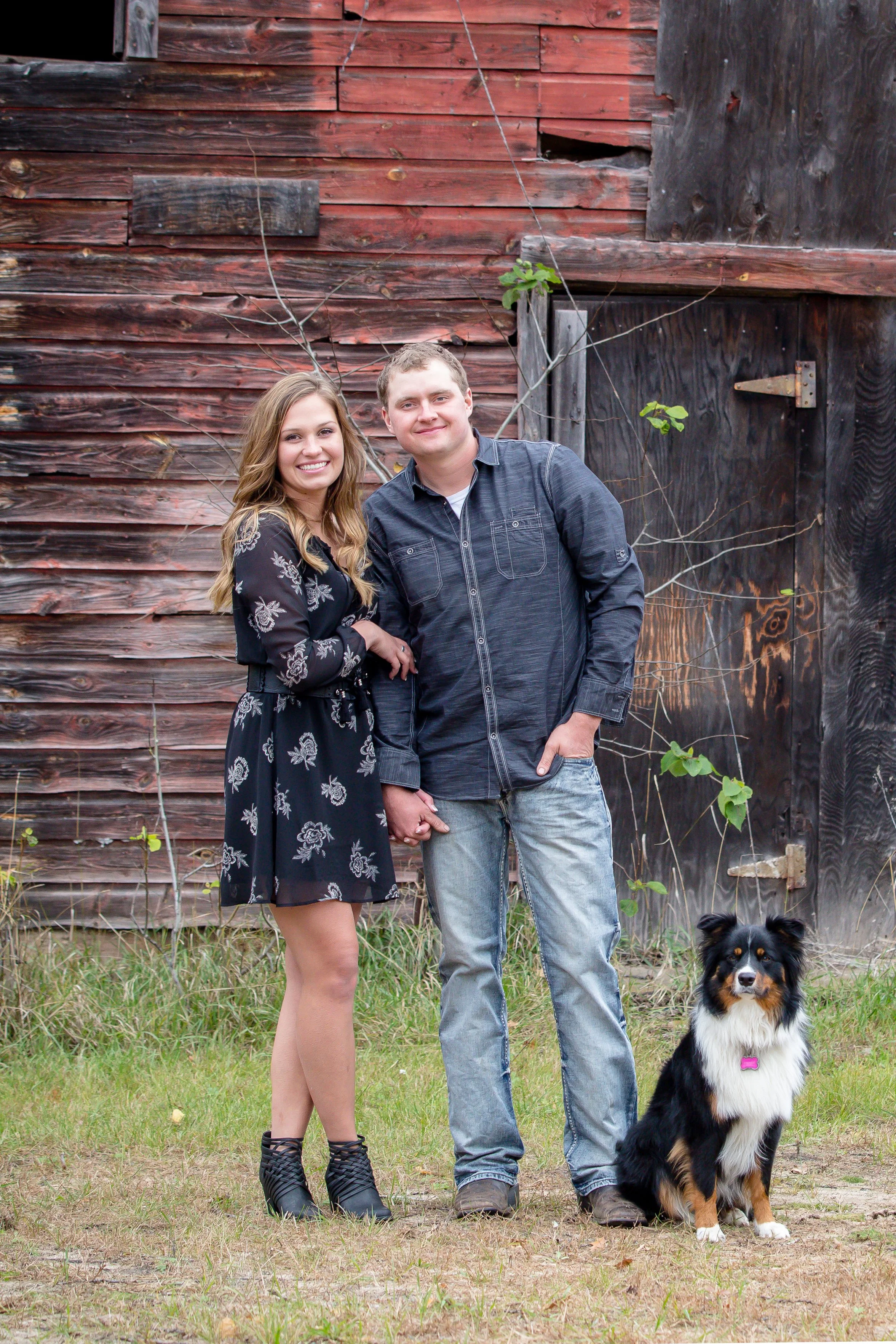A smiling couple stands outdoors holding hands, with a black and white dog sitting beside them in front of a weathered red barn with some small green leaves and vines, taken by MN photographer LKW Photography.