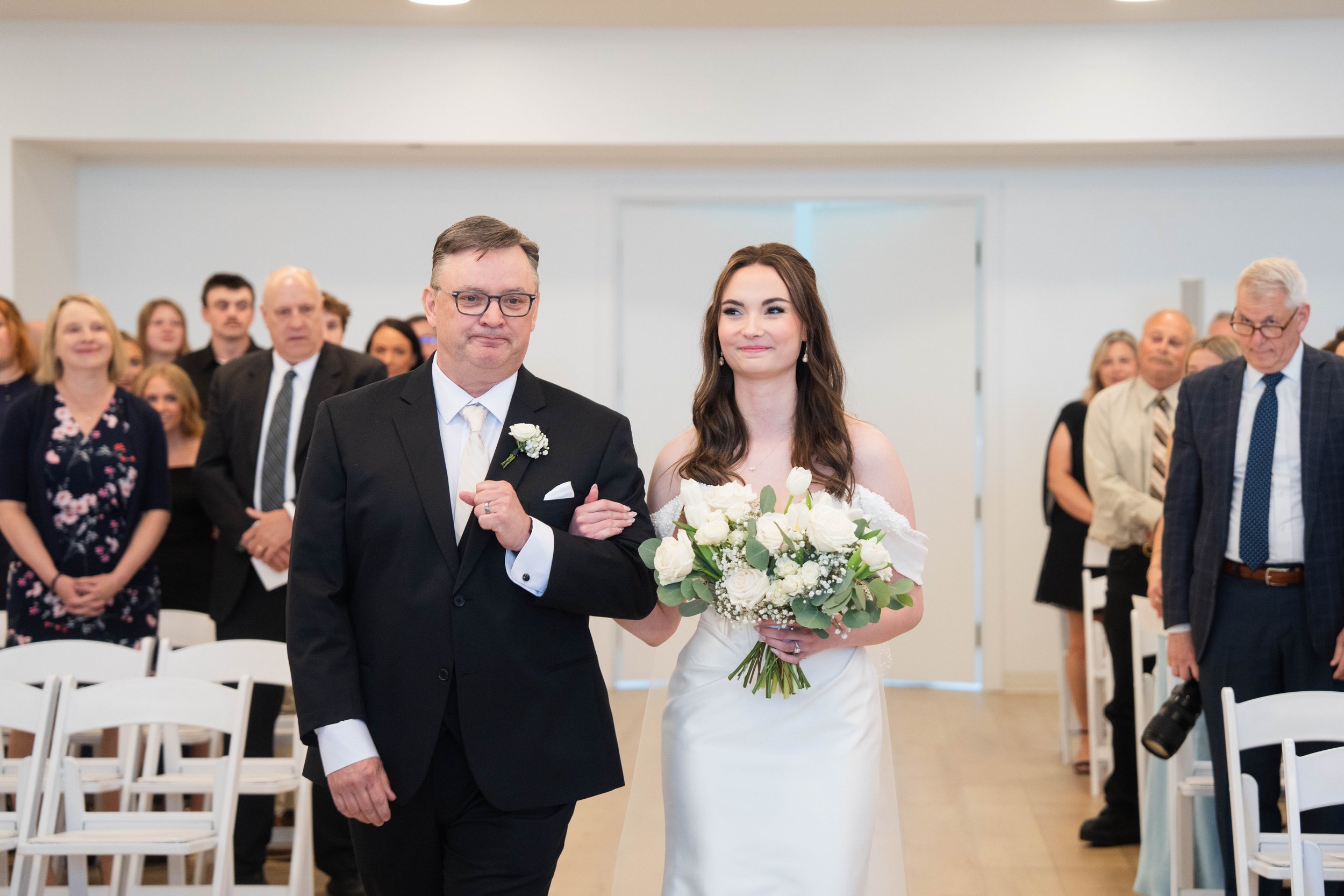 A dad walking his daughter down the aisle on her wedding day, with guests seated and standing in the background.