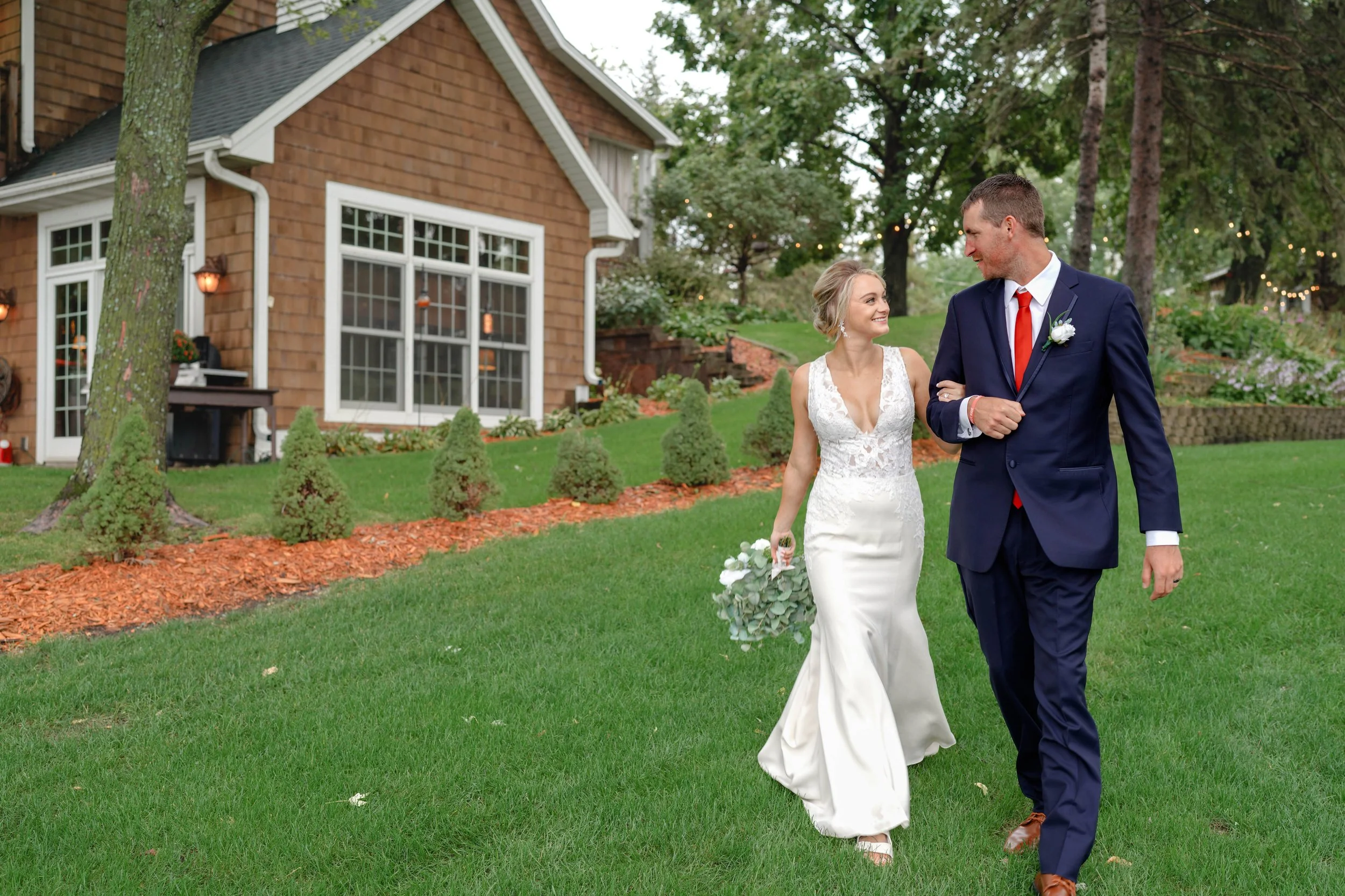 A bride and groom walk in a garden outside a brick house during a wedding. The bride wears a white lace gown and holds a bouquet, and the groom wears a navy suit with a red tie and boutonniere. They smile at each other.