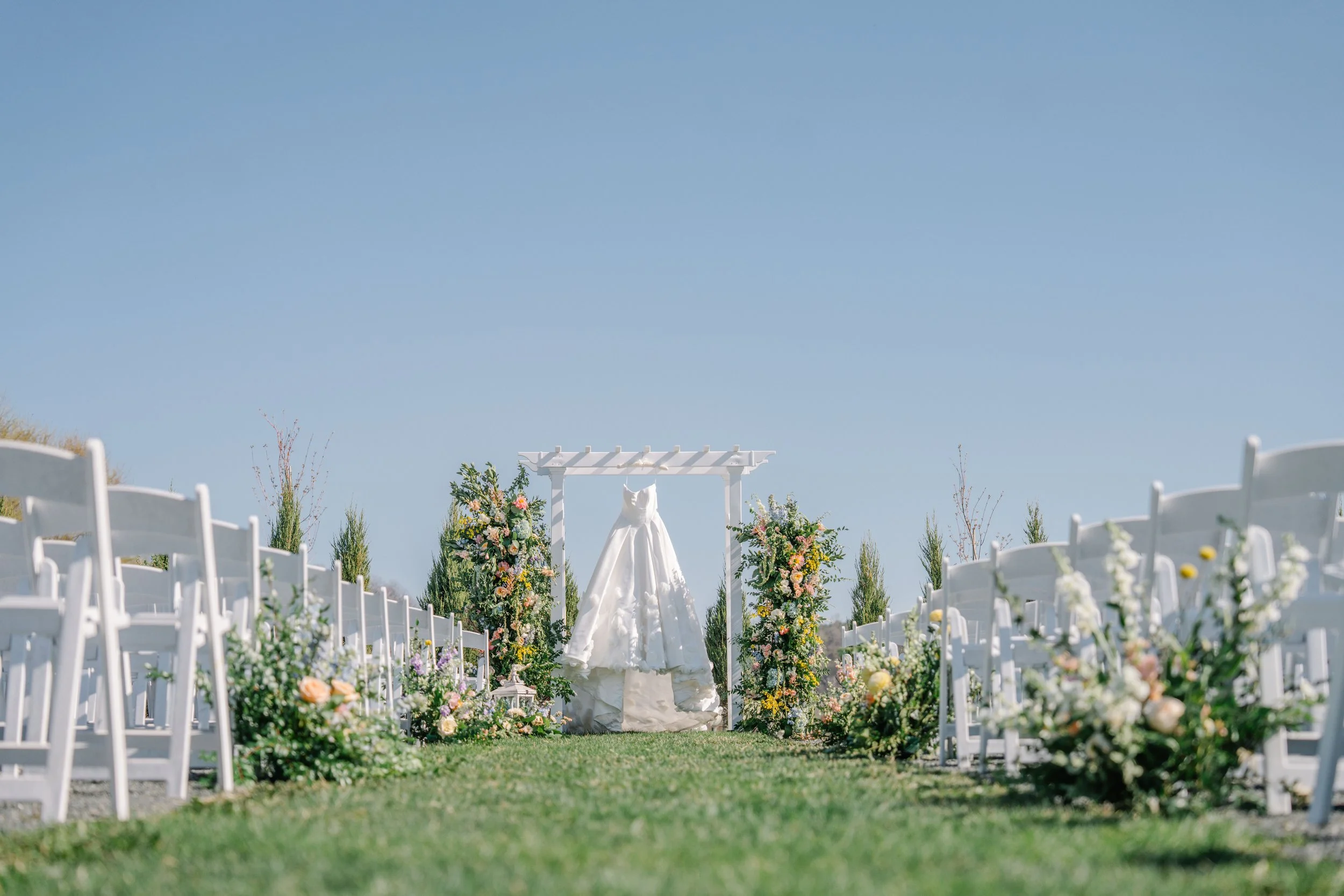 A wedding dress hanging on a trellis decorated with flowers, surrounded by white chairs on a grassy lawn, under a clear blue sky.