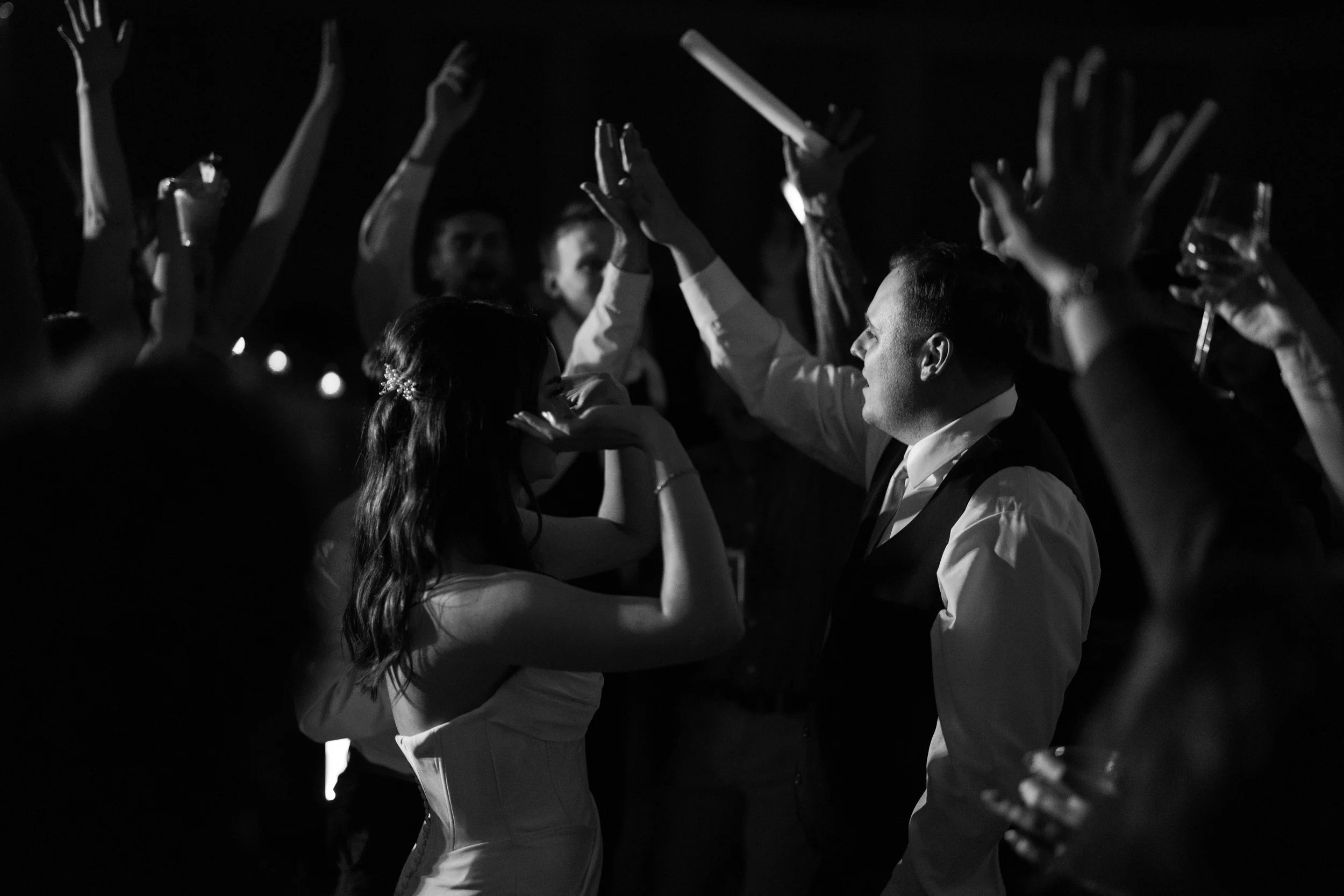 Black and white photo of people dancing at a party, with hands raised in the air, some holding drinks, and a couple in the foreground facing each other.