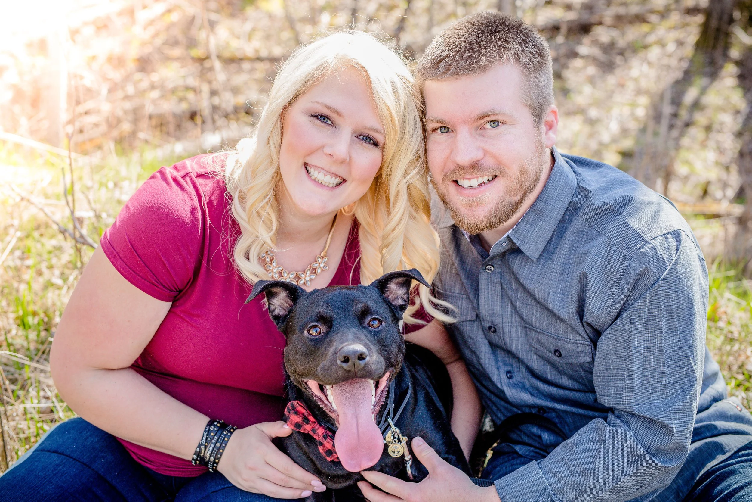 A smiling couple with a black dog outdoors on a sunny day, with trees and grass in the background, taken by Minnesota wedding photographer, LKW Photography.