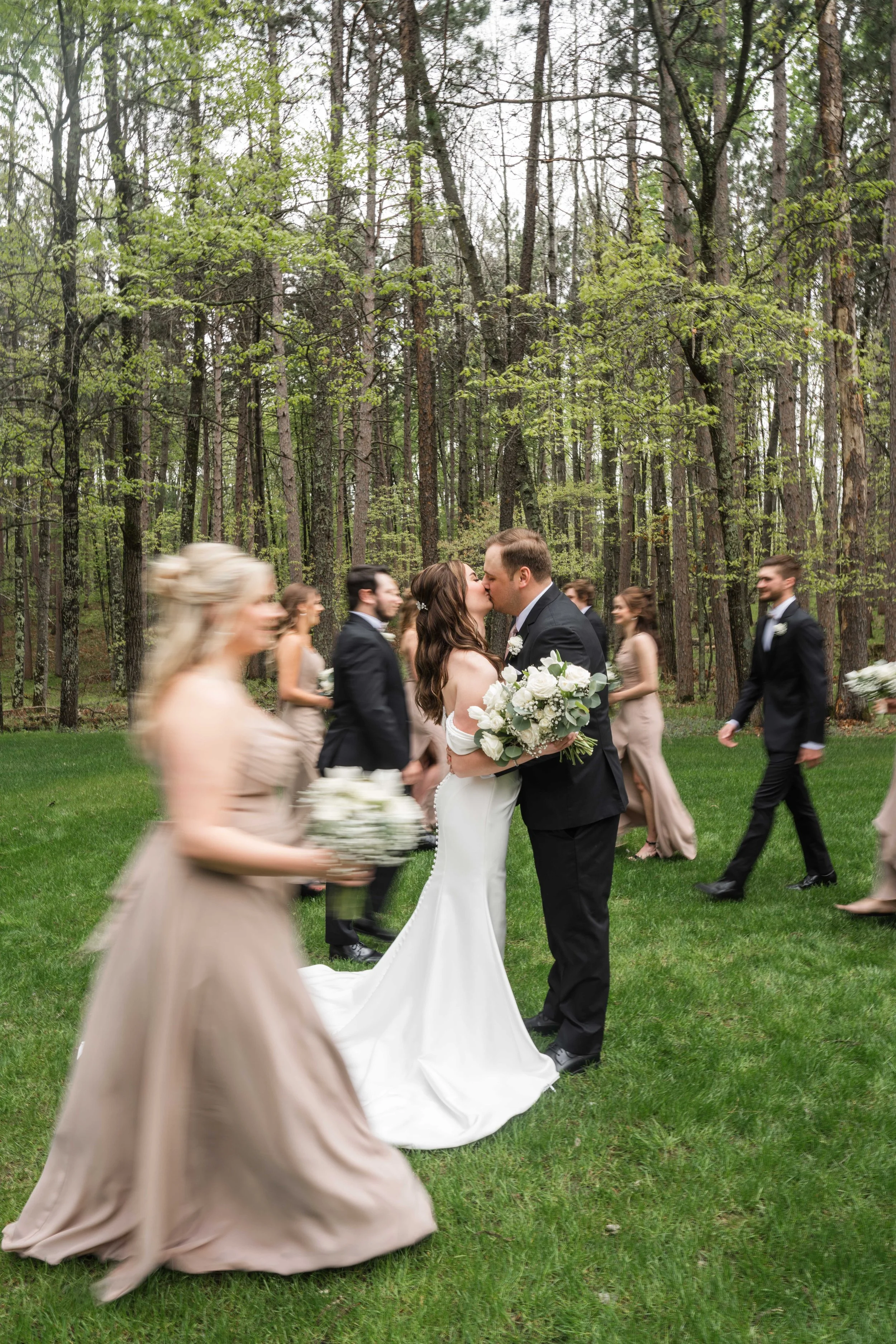 A newlywed couple kisses while walking outdoors on grass, surrounded by wedding guests and tall trees in a forest setting.