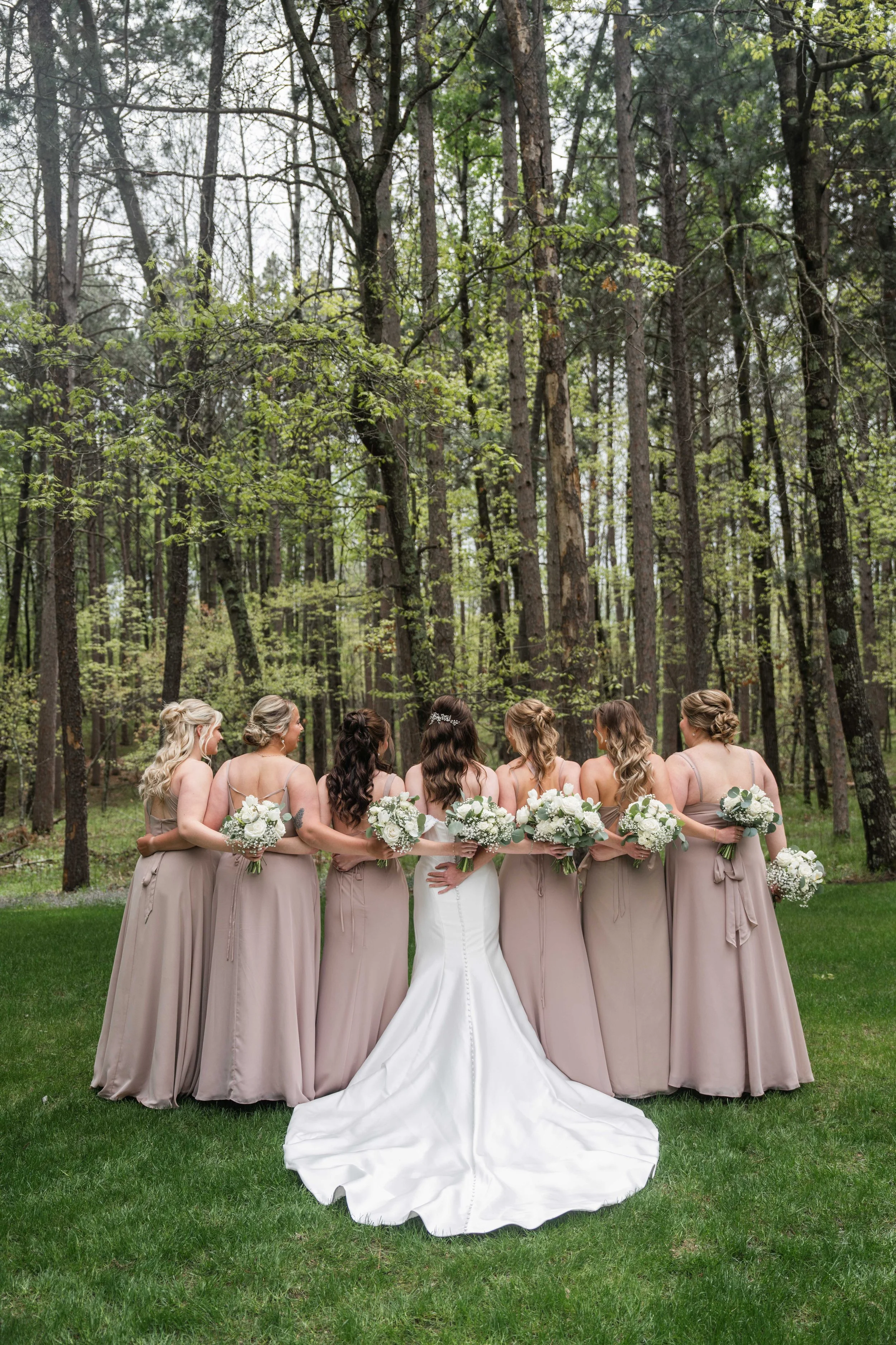 A group of nine women in beige and white wedding dresses, standing outdoors on a grassy area surrounded by tall trees. The women are holding bouquets of white flowers and are viewed from behind.