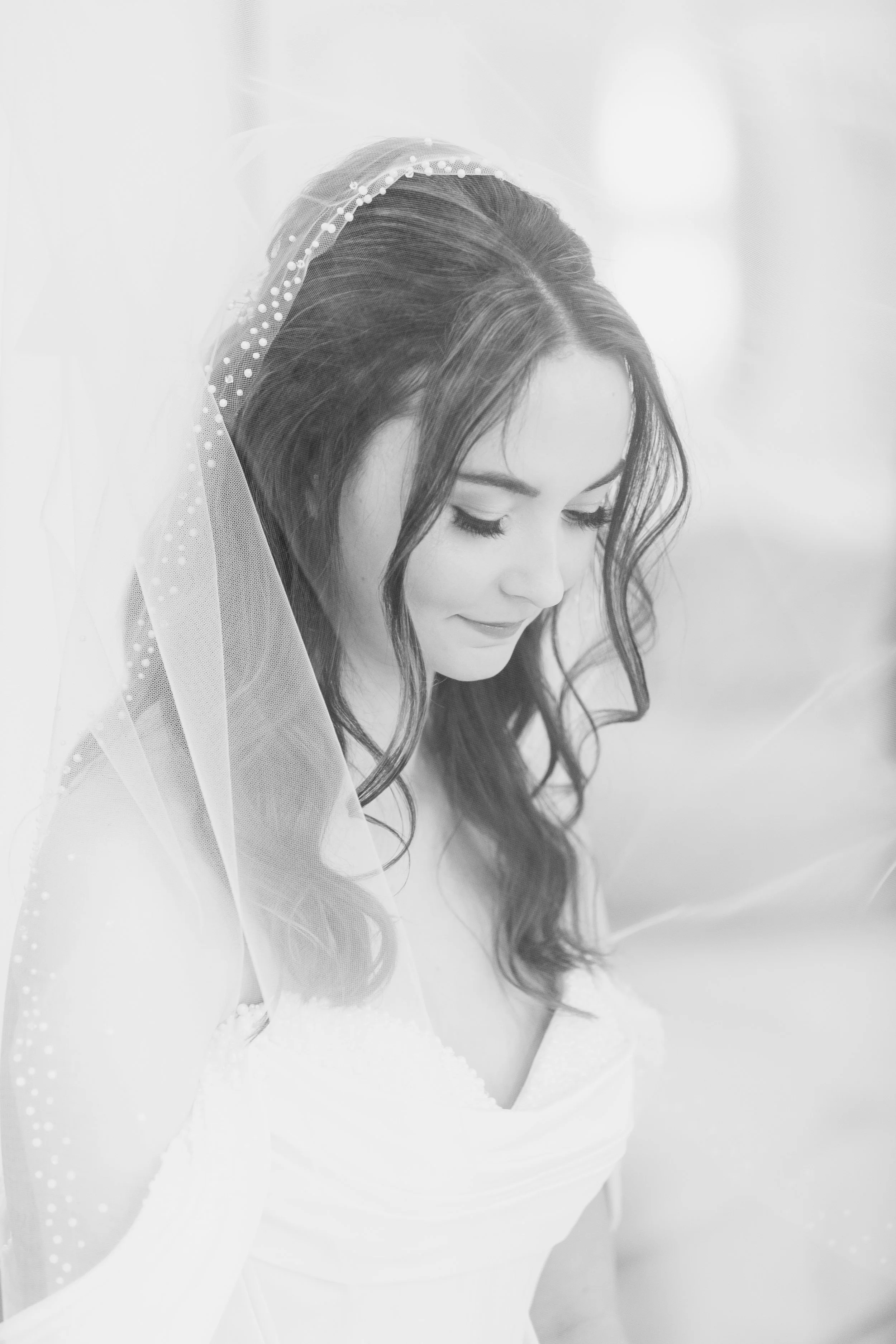 Black and white photo of a bride with long dark hair, wearing a veil with pearls, looking downward, smiling softly.