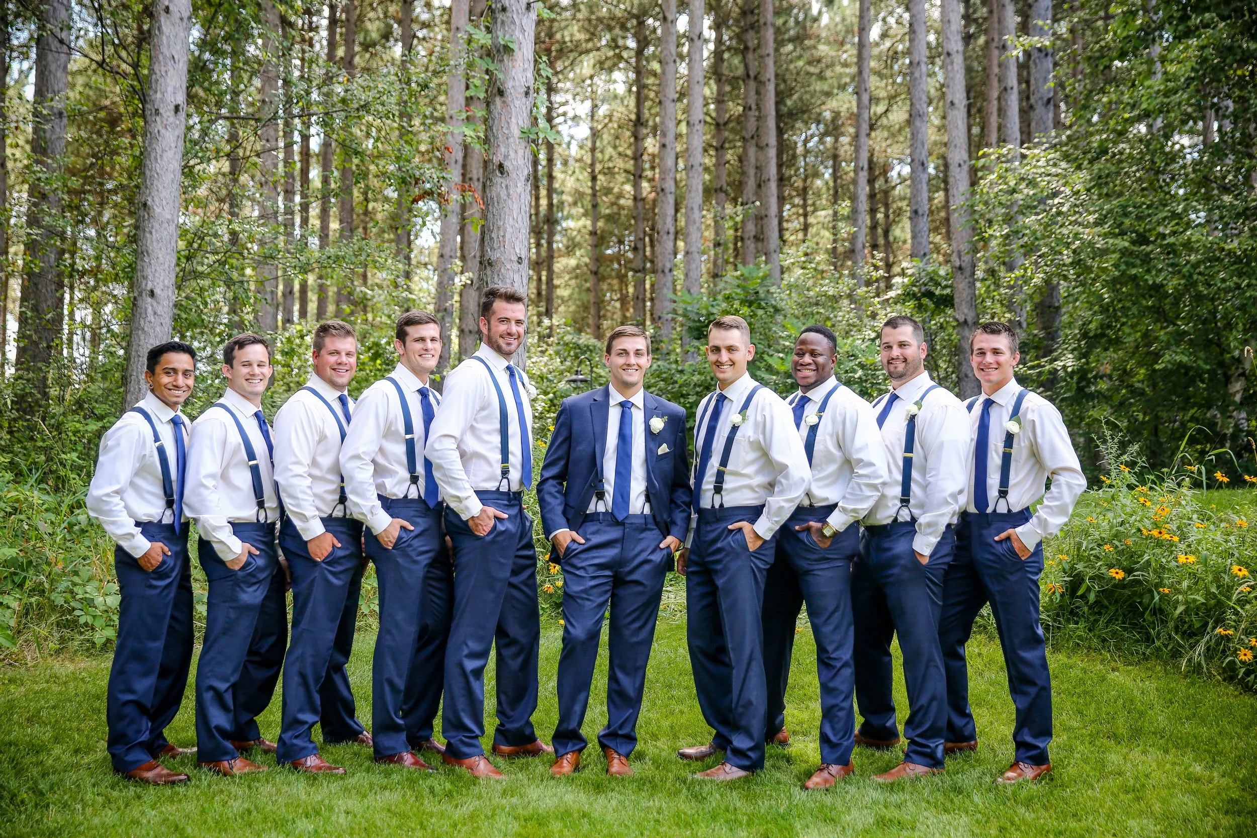 Group of eleven men dressed in formal attire, standing outdoors in a lush, green forest with tall trees and yellow flowers, smiling and posing for a photo taken by Minnesota based wedding photographer, LKW Photography.