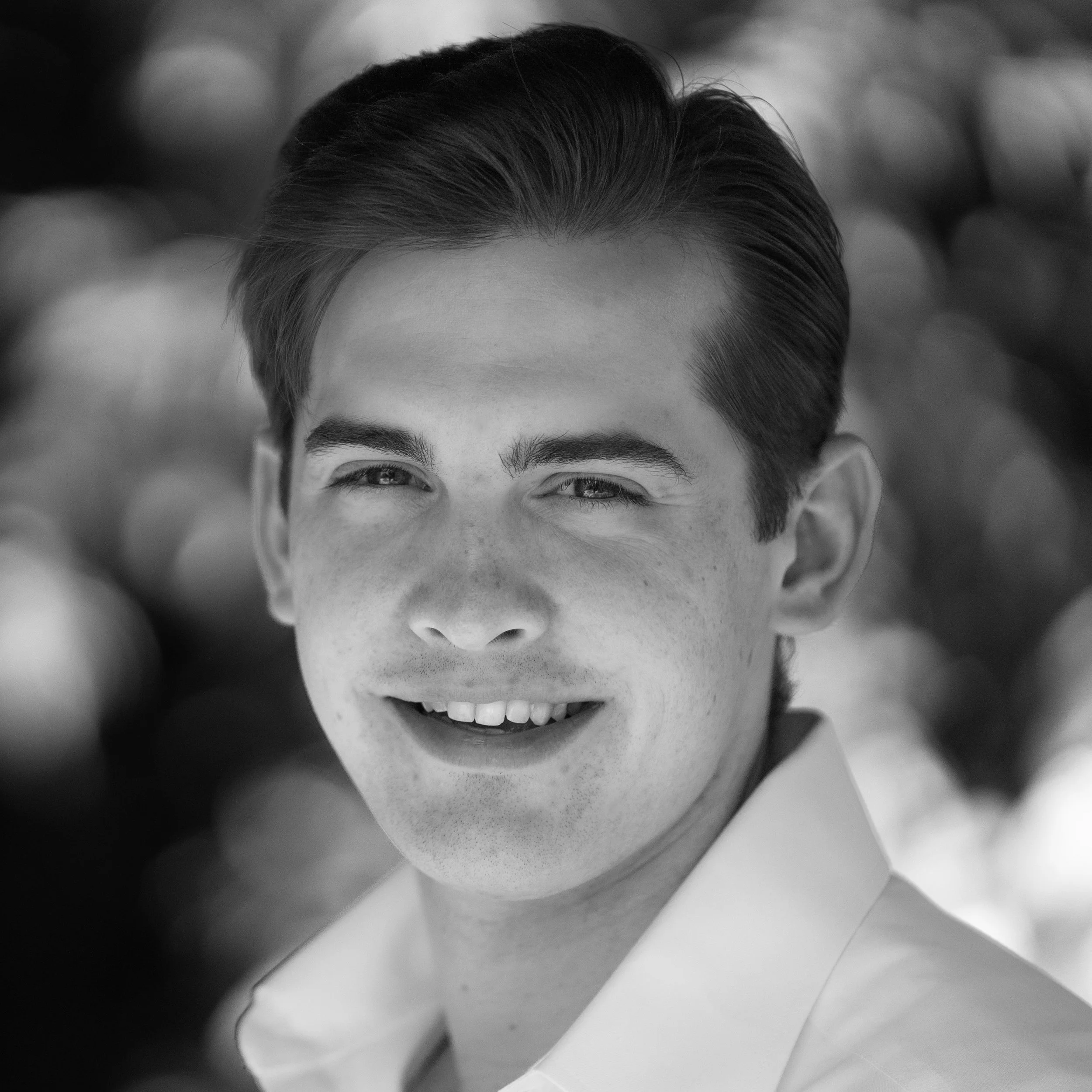 Black and white close-up photo of a young man with neatly styled hair, smiling slightly with a dimple on his cheek, wearing a collared shirt, outdoors with blurred foliage in the background.