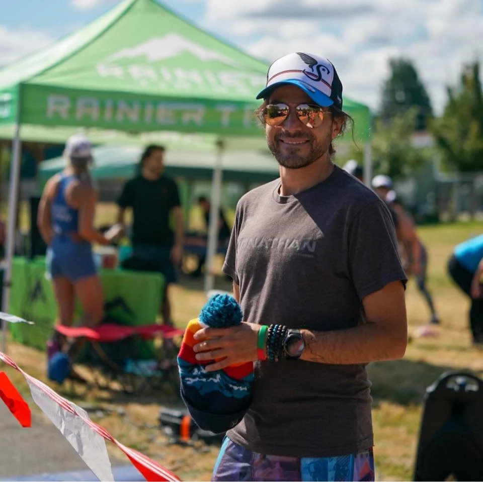 Smiling man wearing sunglasses, a cap, a gray T-shirt, and colorful shorts holds a colorful beanie hat with a pom-pom at an outdoor event with green tents and other people in the background.