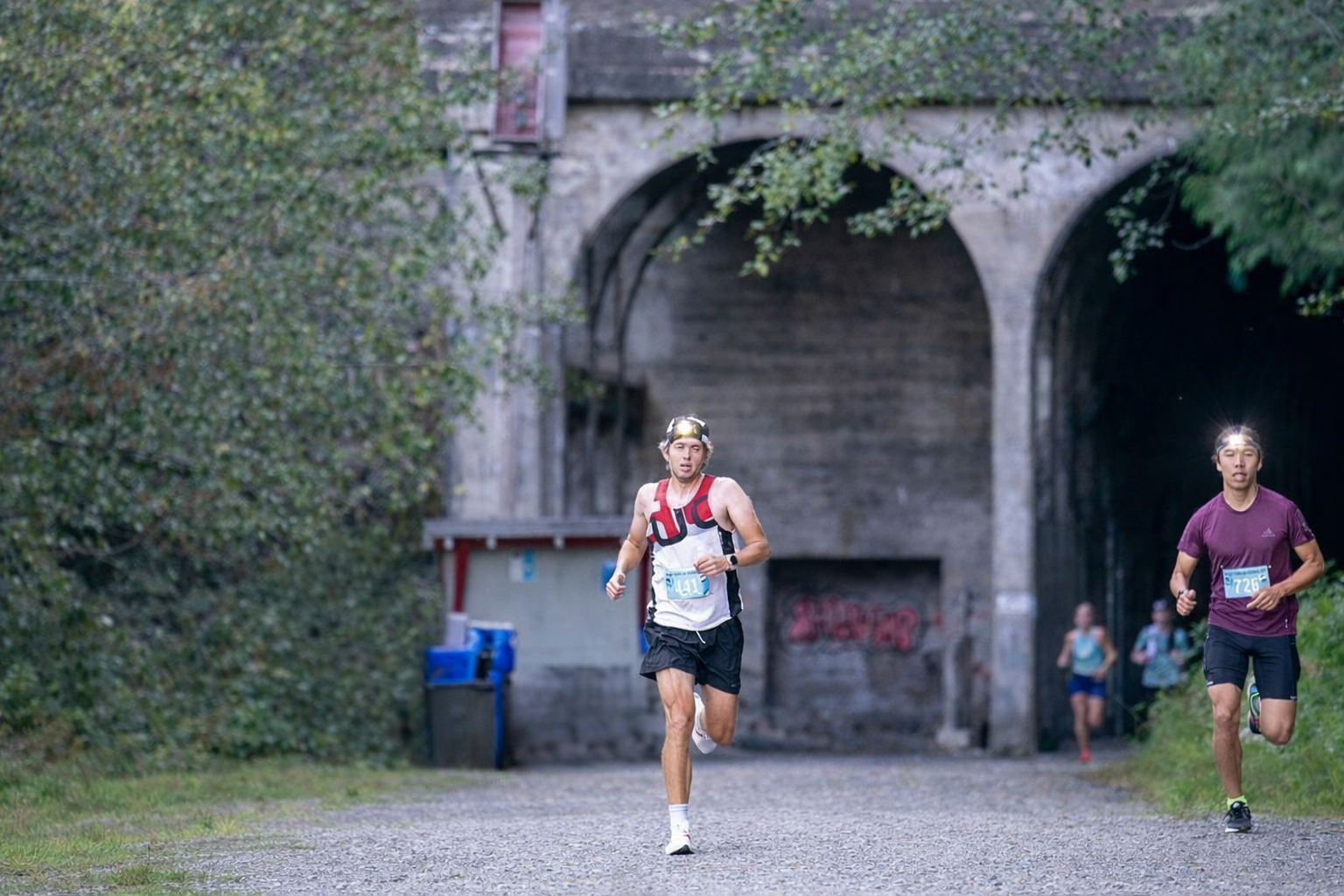 Two runners participating in a race on a gravel trail near a tunnel, surrounded by greenery.