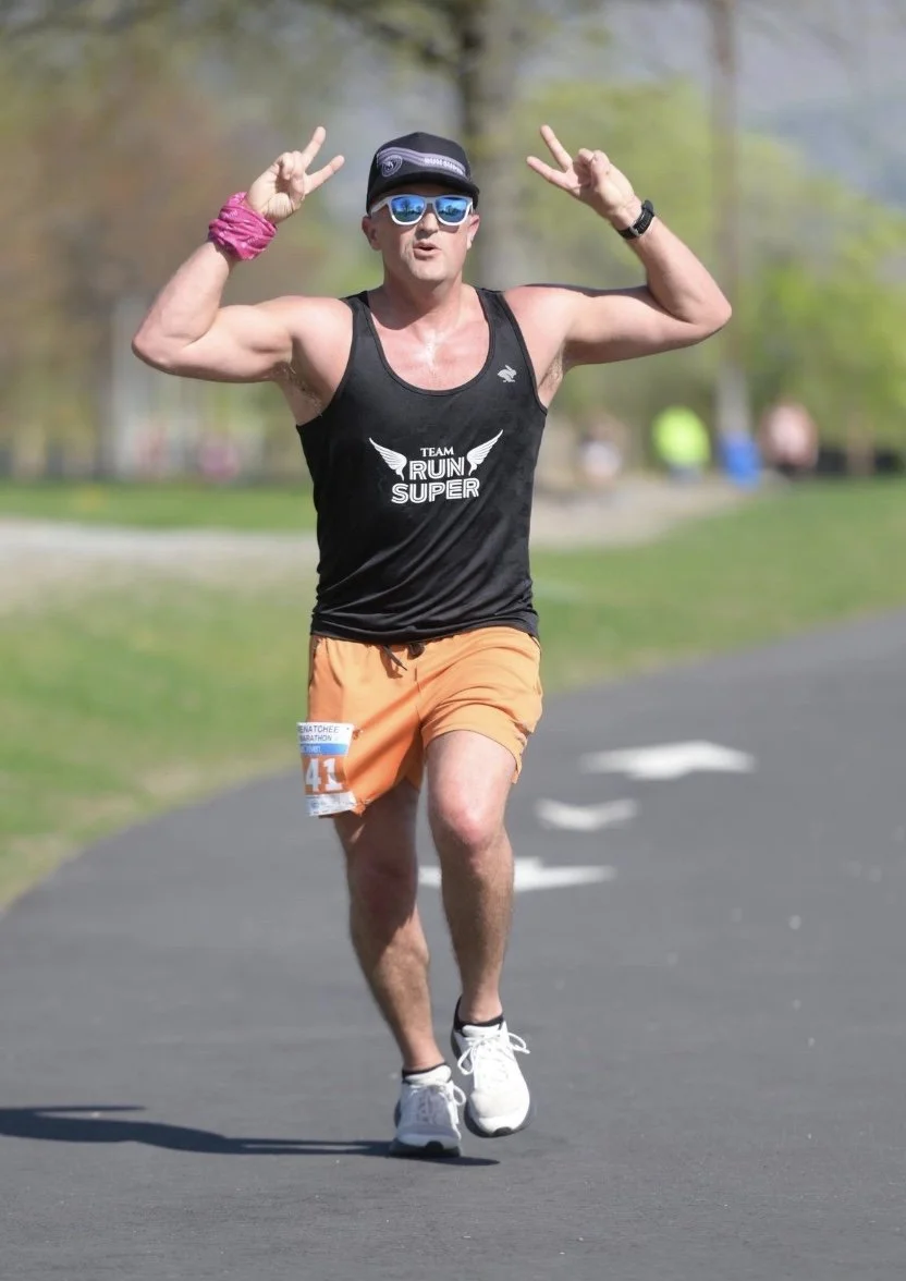 A man running in a marathon, wearing a black tank top with 'TEAM RUN SUPER' written on it, orange shorts, sunglasses, a black cap, and making peace signs with both hands.