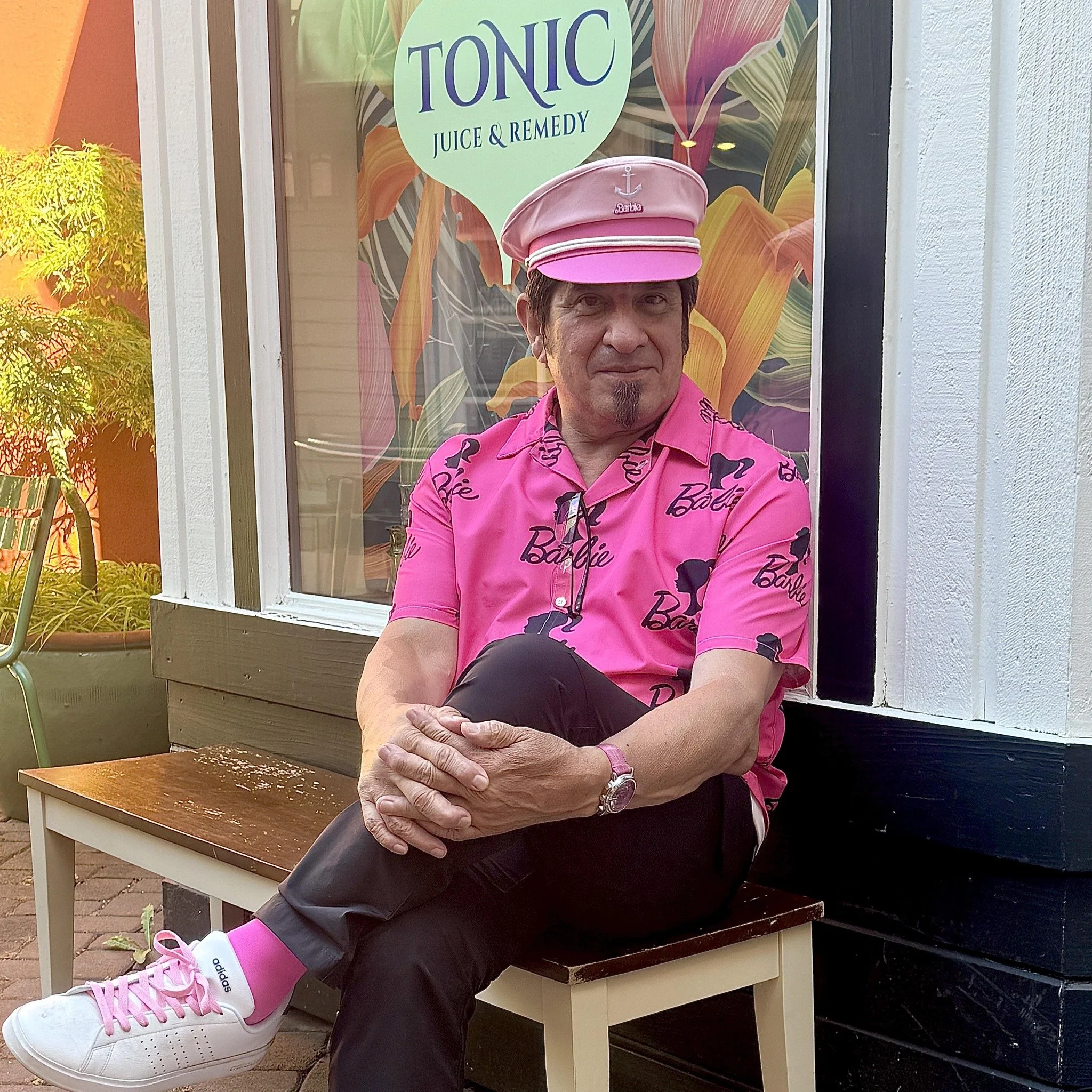 A man sitting on a bench outside a storefront with a colorful floral window display. He's wearing a pink Barbie themed shirt, matching pink hat, and white sneakers with pink socks. The storefront sign reads "Tonic Juice & Remedy."
