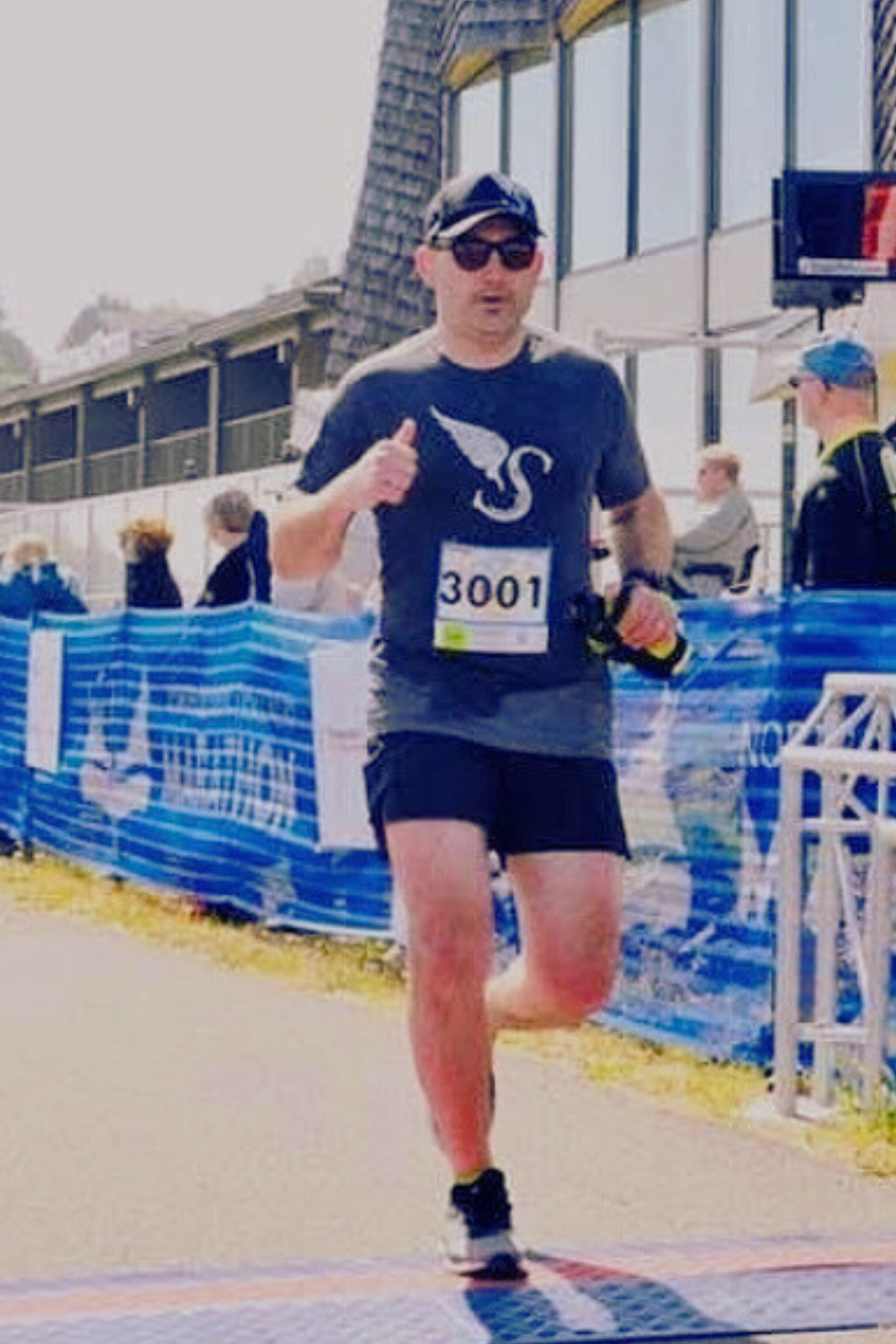 A male runner wearing sunglasses, a black t-shirt with a white swan logo, black shorts, a cap, and a race bib number 3001, running outdoors near a blue barrier with people and modern buildings in the background.