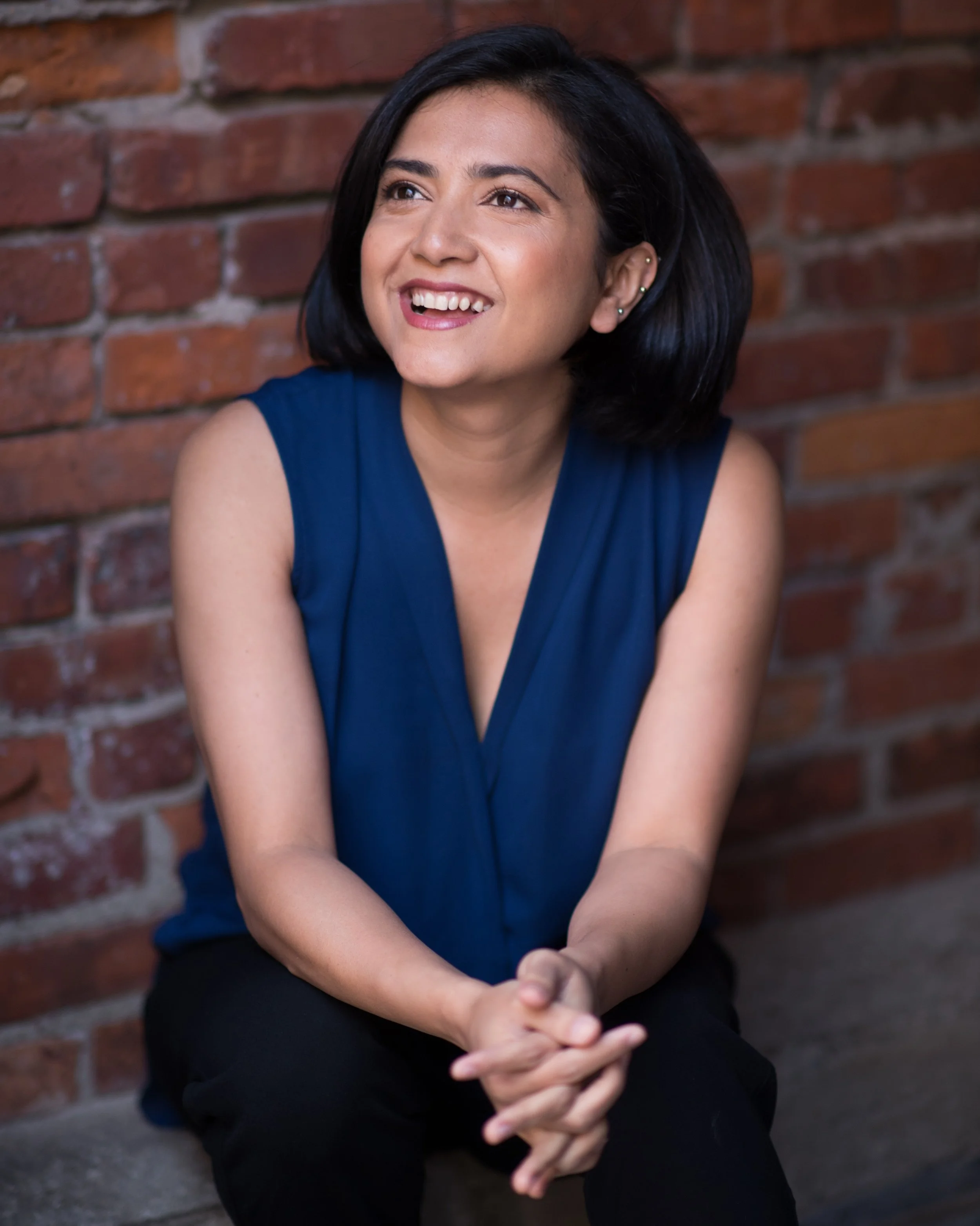 A woman with short dark hair, wearing a sleeveless blue top, sitting against a brick wall with her hands clasped. She is smiling and looking slightly to her right.