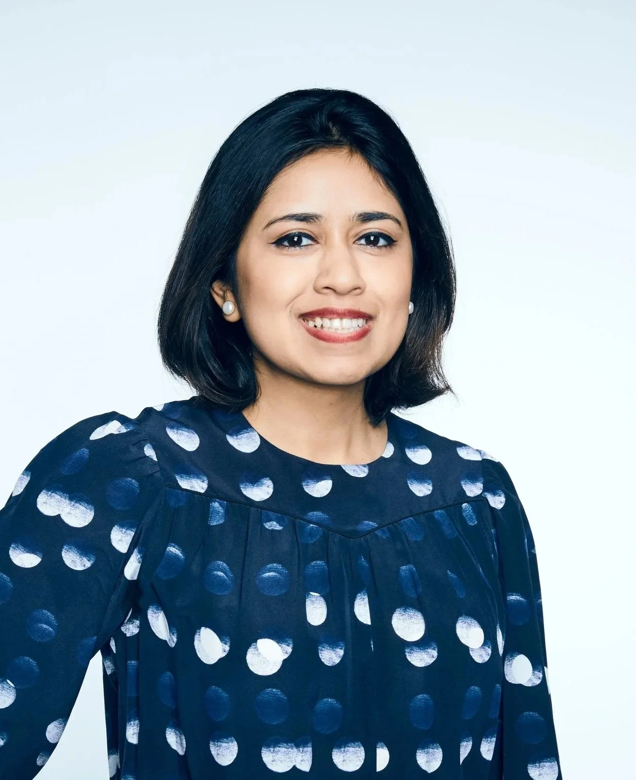 A woman with shoulder-length black hair, dressed in a navy blue polka dot dress and pearl earrings, smiling in front of a plain light-colored background.