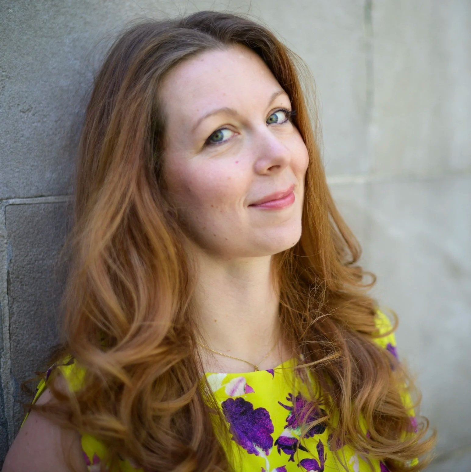 A woman with long, curly red hair leaning against a gray stone wall, wearing a yellow dress with purple and green floral patterns.