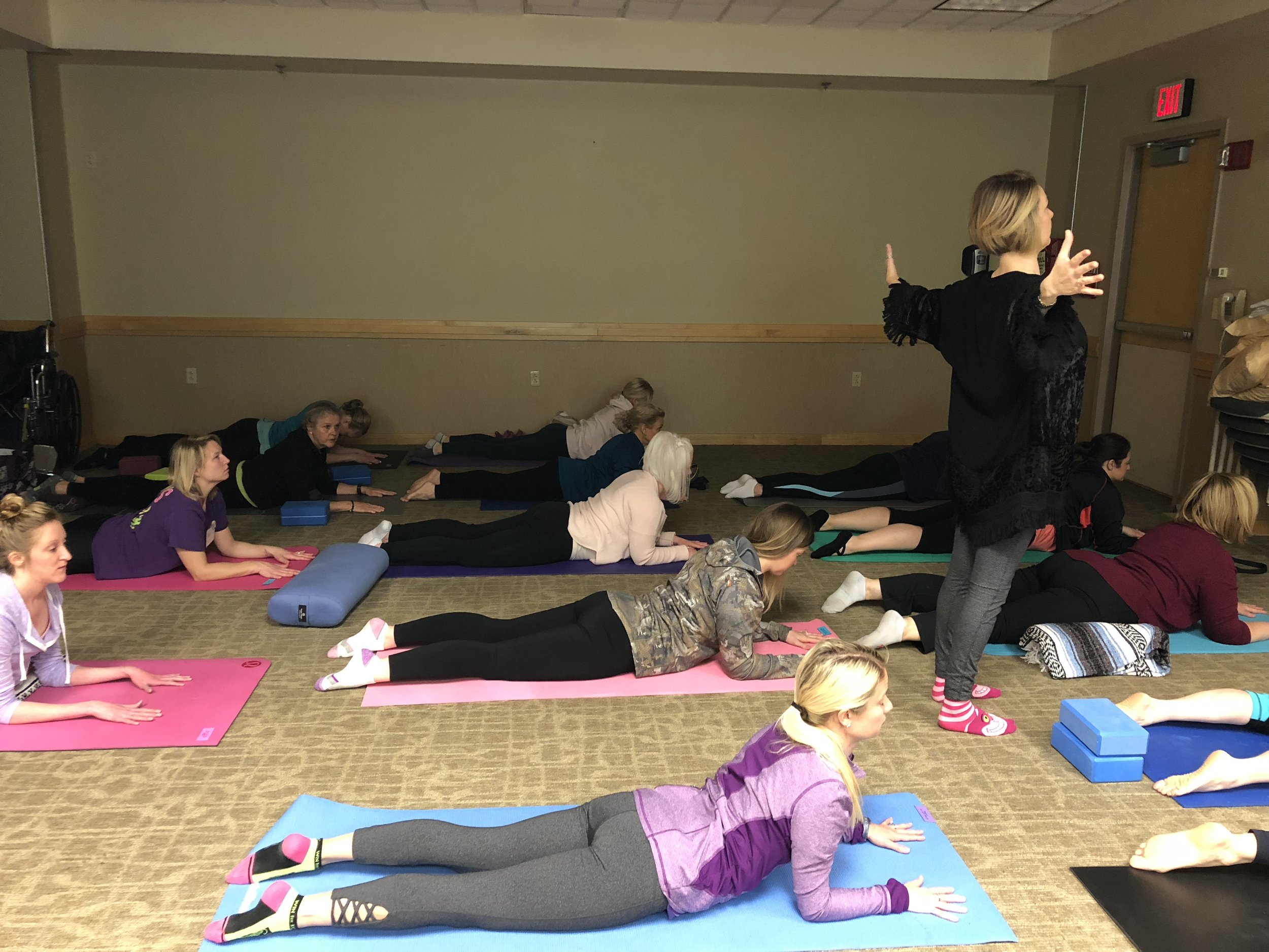 A group of women and girls participating in a yoga class, lying on colorful mats on the floor with a woman in the foreground leading the class in a pose with arms extended through a doorway.