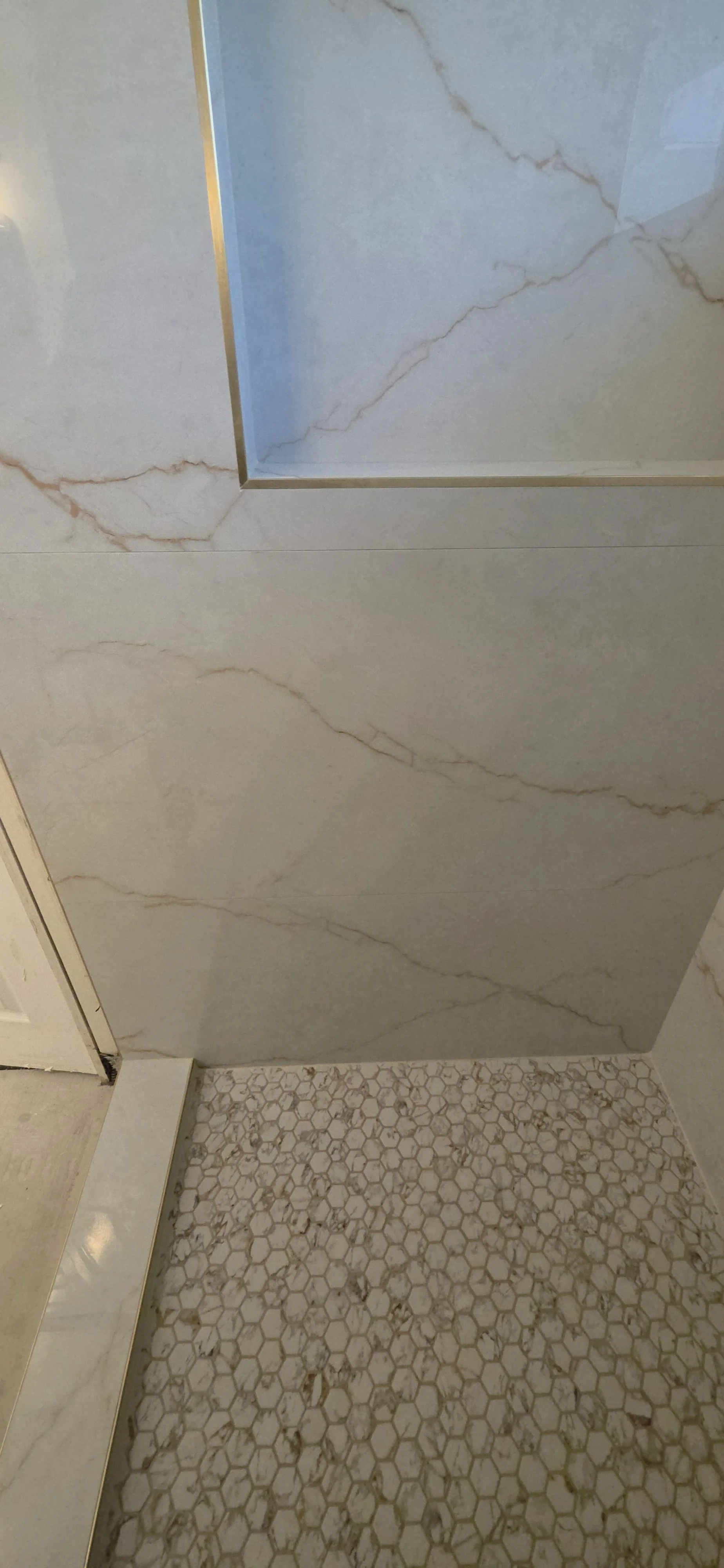 Mosaic shower floor and tiled curb beneath marble-look wall tile, with gold-tone trim at the recessed niche. Tilework by Arroyos Tile and Granite Inc.; remaining fixtures and bathroom finishes are pending.
