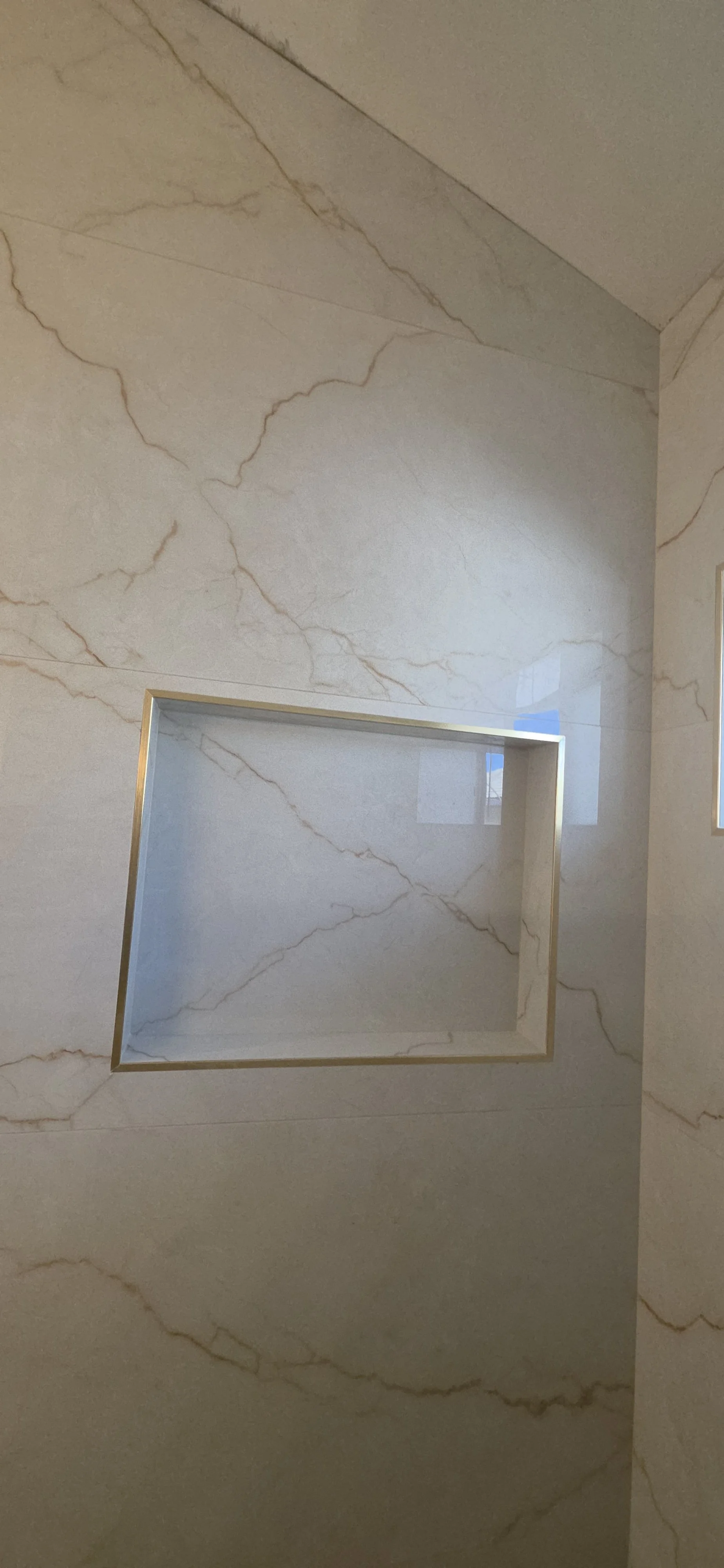 Recessed shower niche with gold-tone trim set into marble-look wall tile. Niche and tile detail by Arroyos Tile and Granite Inc., shown before the remaining bathroom fixtures and finishes are installed.