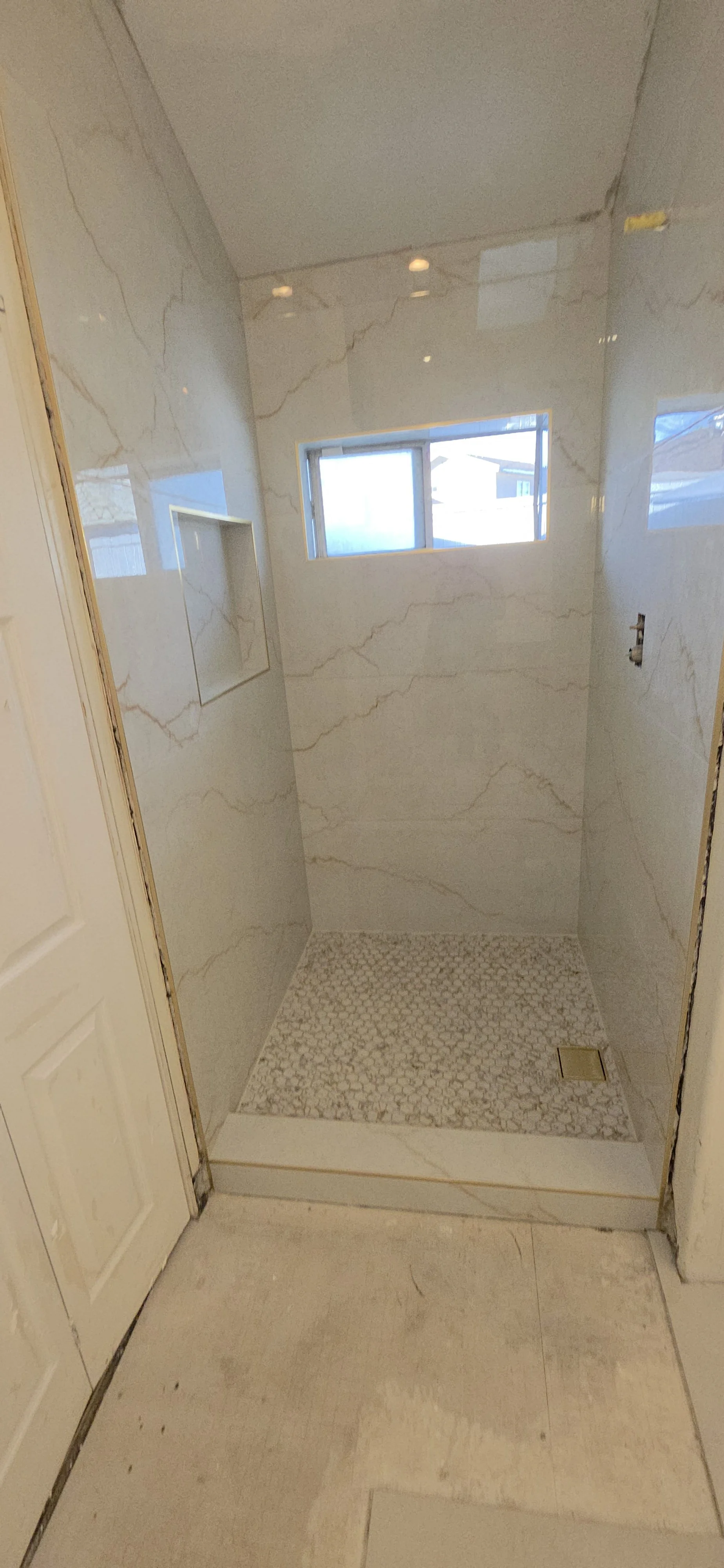Shower tilework by Arroyos Tile and Granite Inc.: marble-look wall tile, a recessed niche with gold-tone trim, a window, mosaic floor and tiled curb. Shown before the remaining fixtures and bathroom finishes are installed.