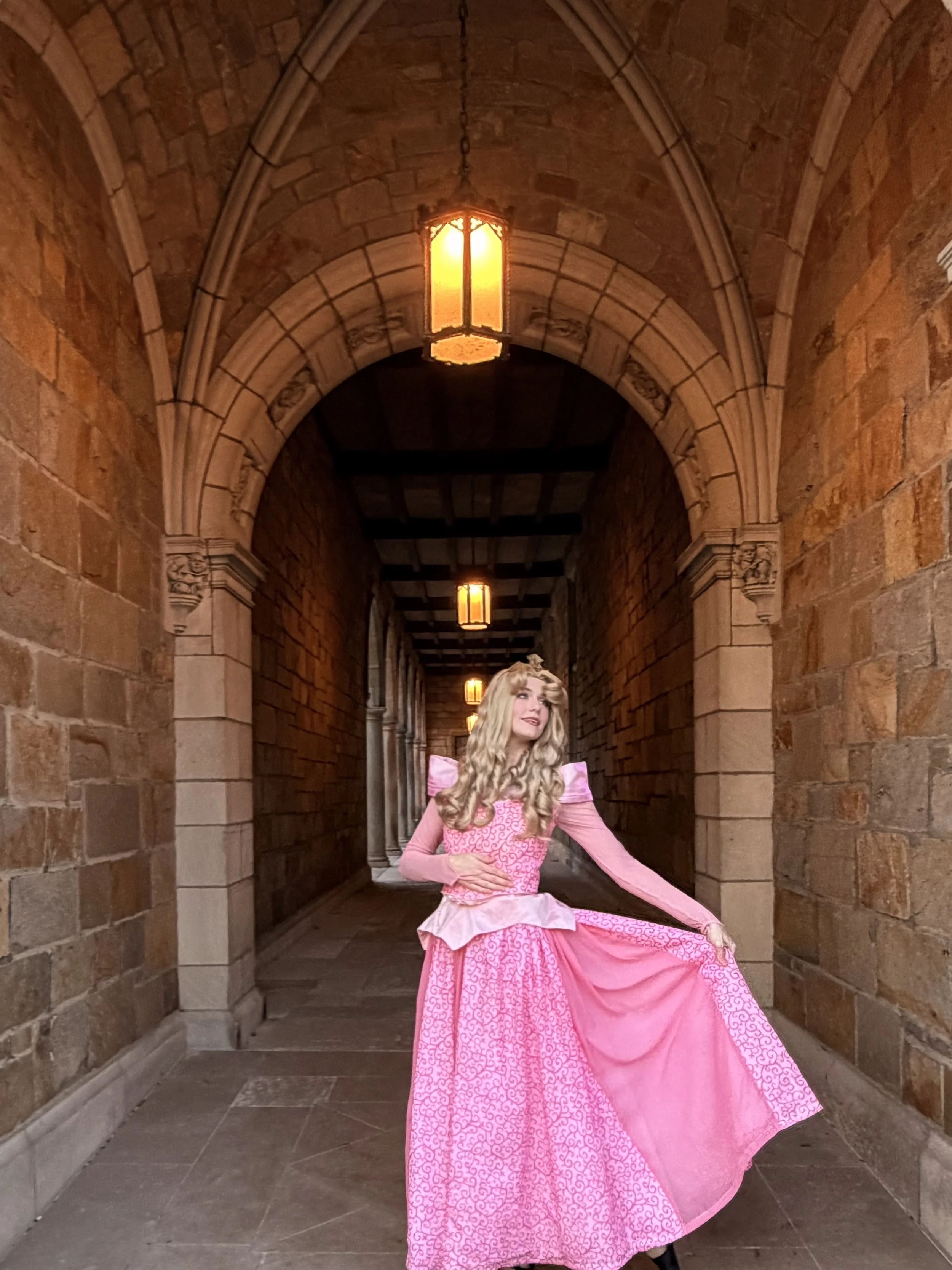 Person in a pink princess costume with a crown, lying on a matching patterned pink fabric, smiling. sleeping beauty face character, aurora face character, aurora birthday party, princess party