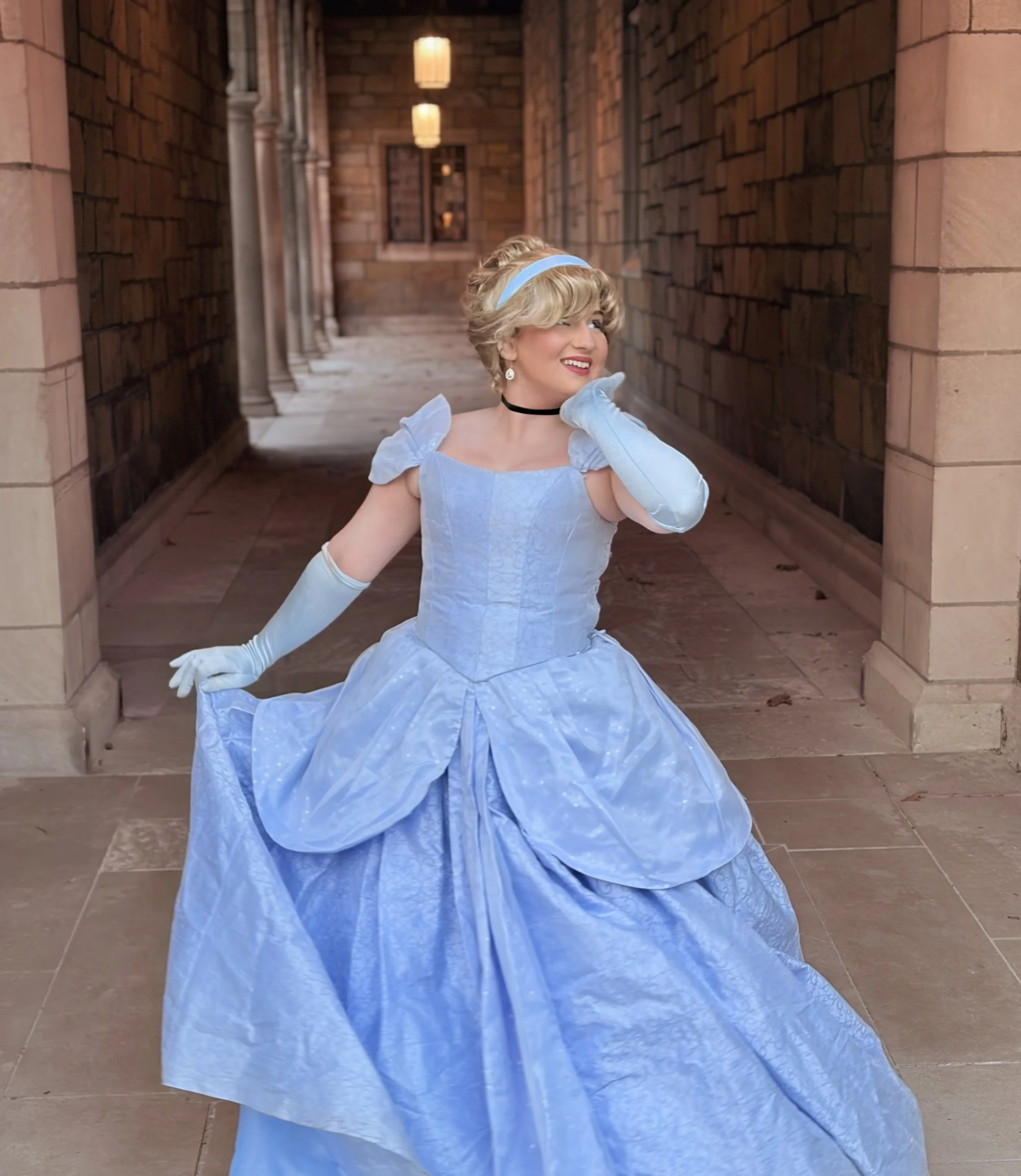 Person dressed as a princess in a light blue gown sitting on stone steps outdoors, smiling and waving. Cinderella  face character, face character, princess birthday party, princess party in Michigan cinderella for hire
