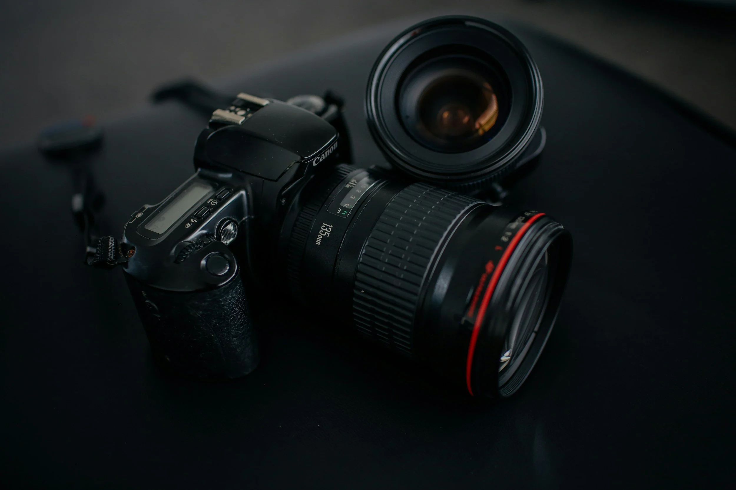 A professional Canon camera with a large zoom lens and an additional lens hood attached, resting on a dark surface.