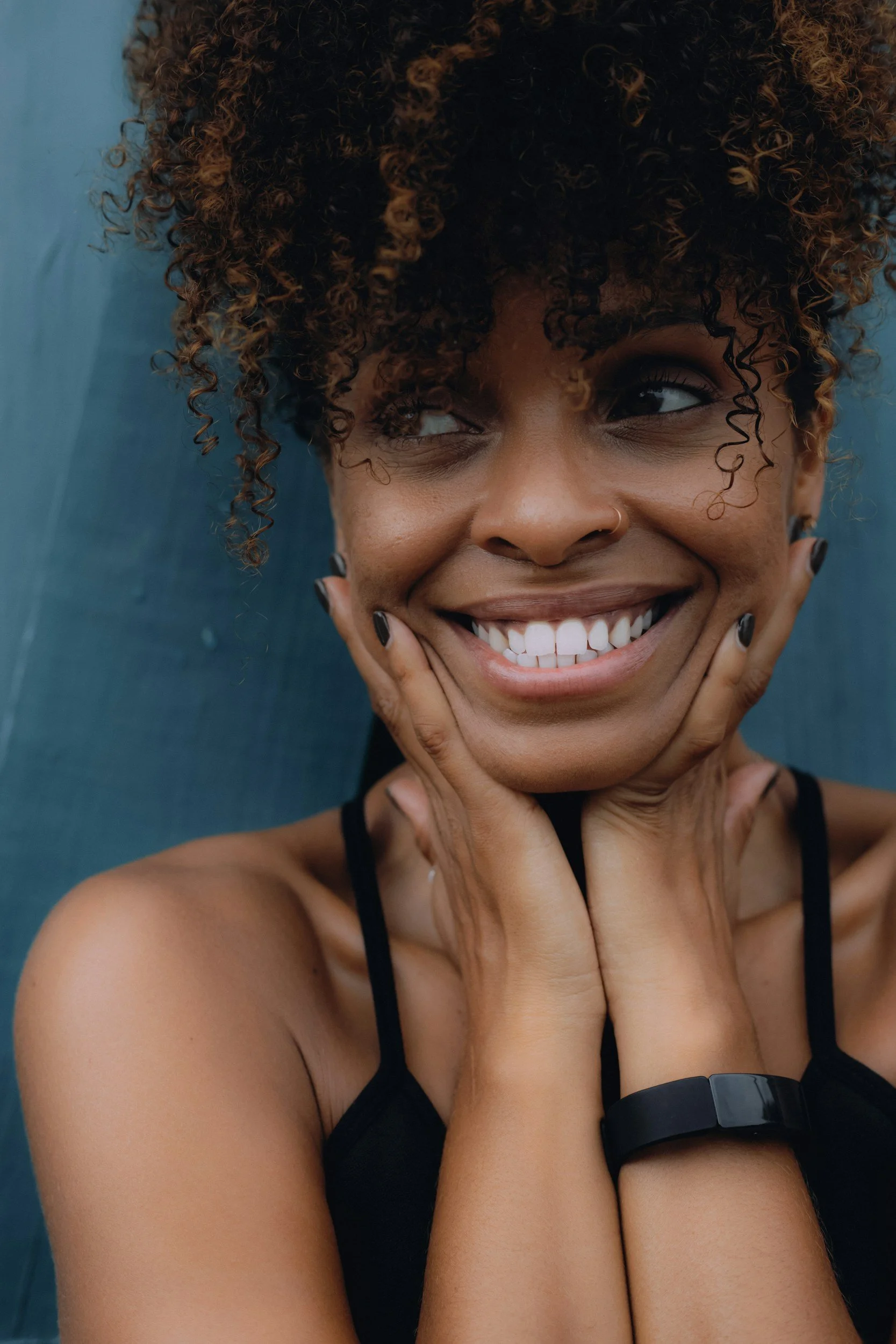A smiling woman with curly hair, wearing a black tank top and a watch, touching her face with her hands, against a blue background.