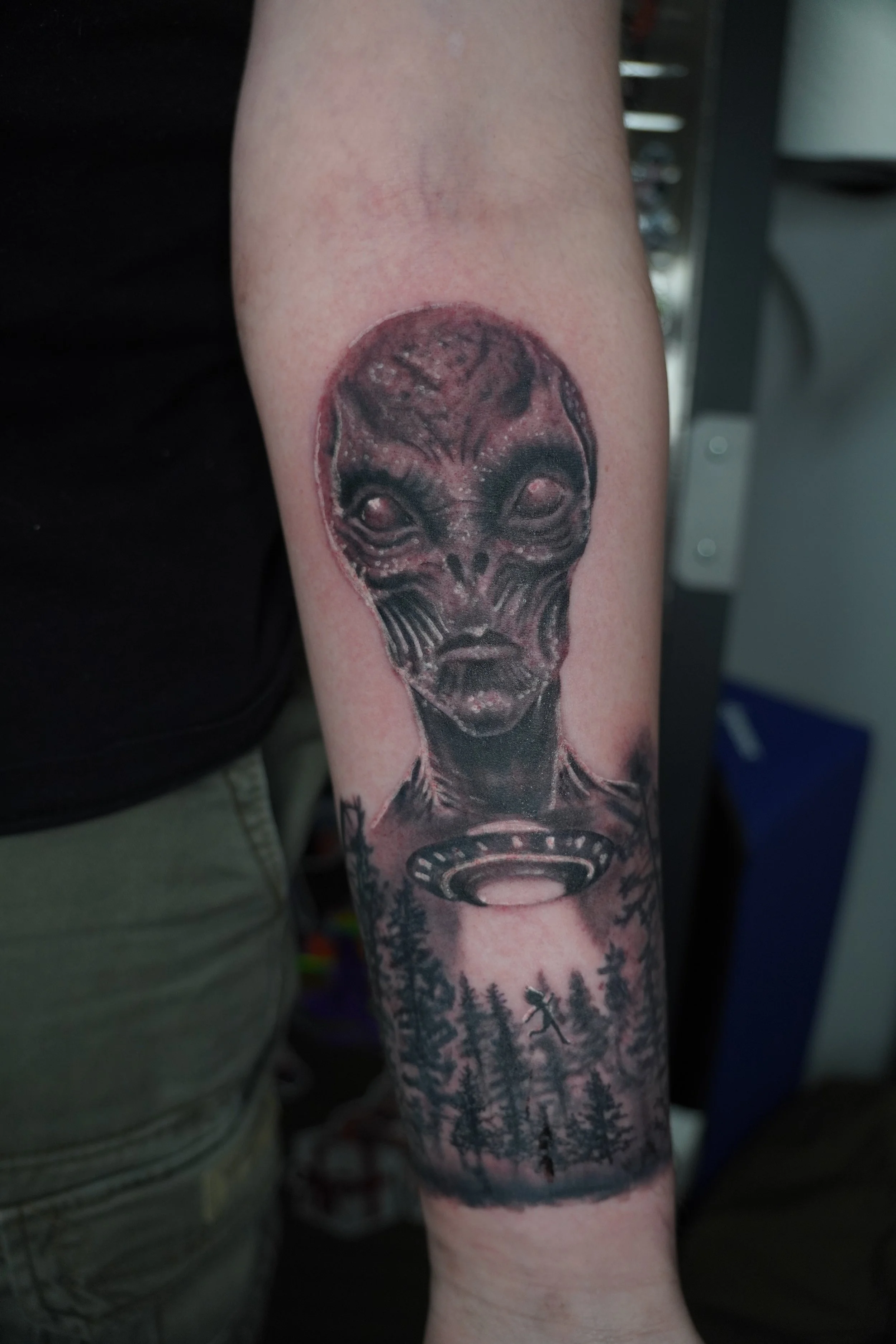 Tattoo of an alien with large eyes and elongated head, flying a UFO over a forest at night.