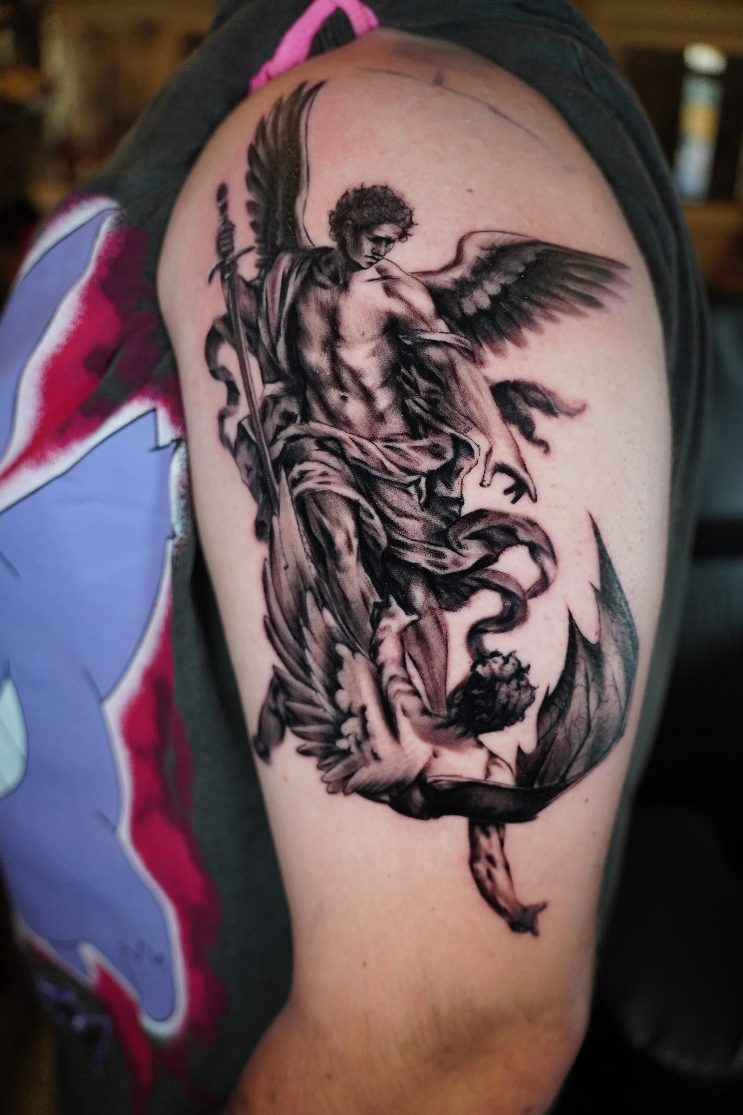 Black and gray tattoo of a winged angel holding a sword, standing over a dragon on upper arm.