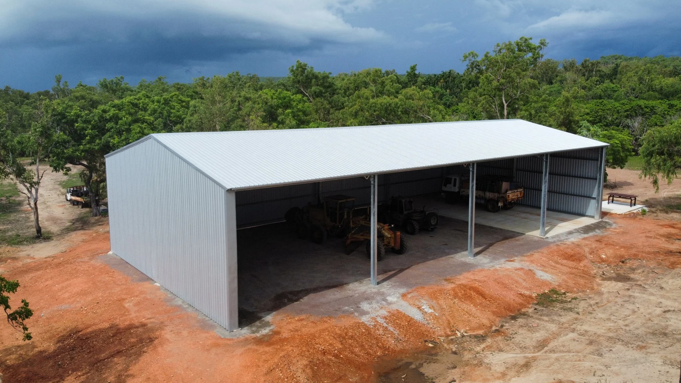 A large metal shed with an open front, containing construction or agricultural vehicles, situated in an open area with trees and a cloudy sky in the background.