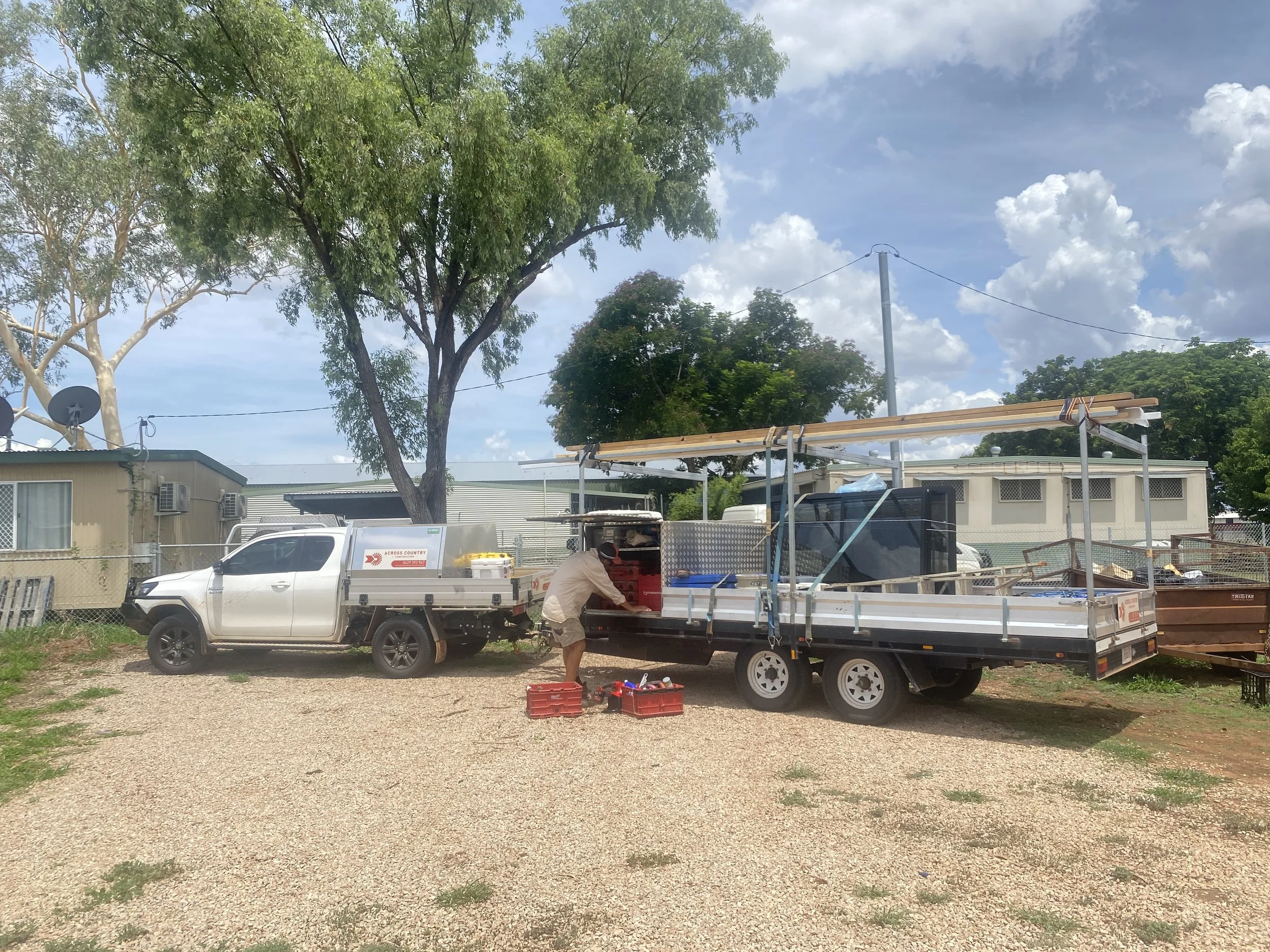 A man loading tools from a red toolbox into a trailer attached to a white pickup truck parked on a gravel lot. The trailer contains wooden planks and other construction materials, with a partially open black mesh cover. There are trees, mobile homes, and a partly cloudy sky in the background.