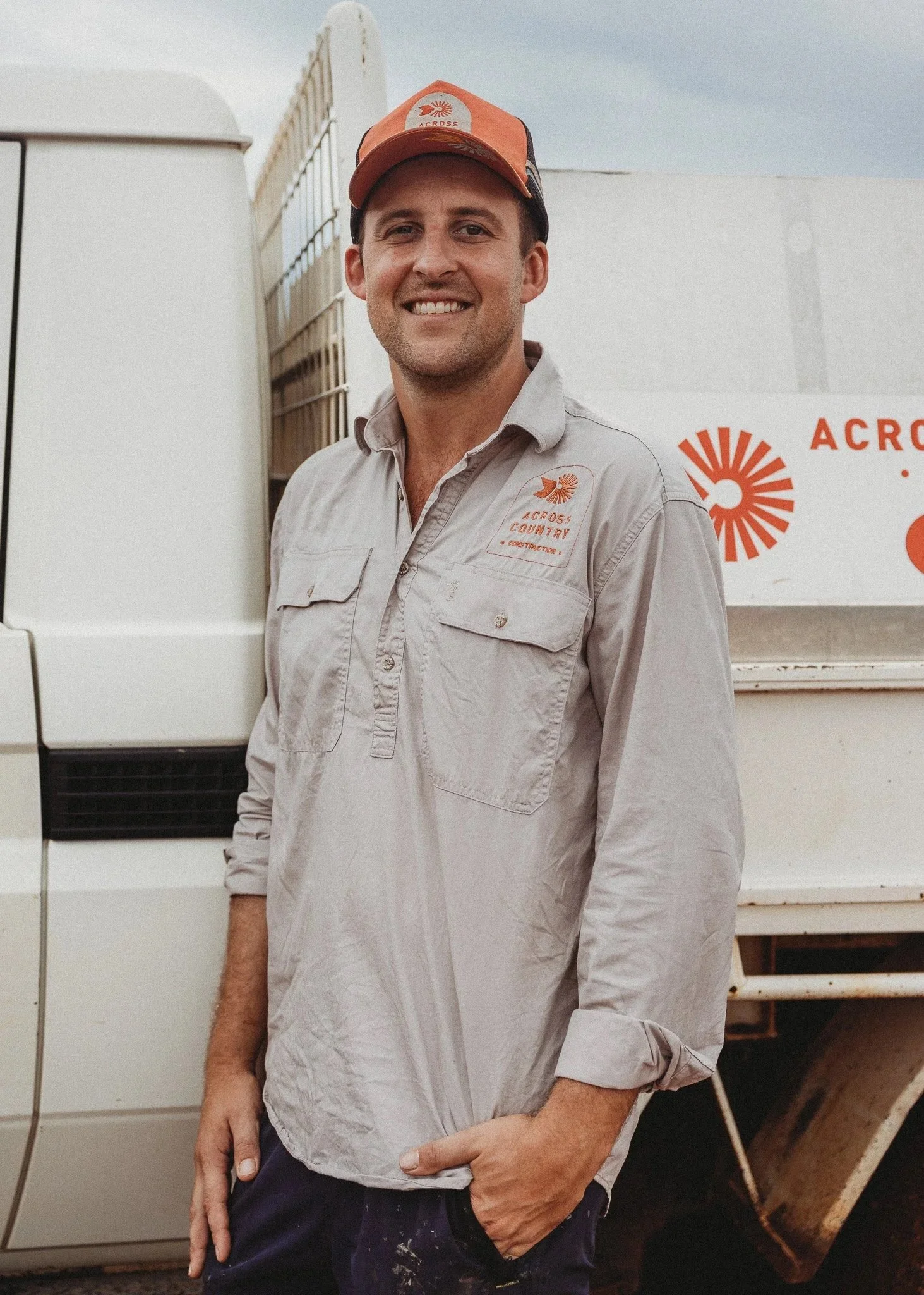 A man smiling outdoors, wearing a light beige work shirt and an orange cap, standing in front of a white truck with a logo and text related to 'Across' and 'Country Construction'.