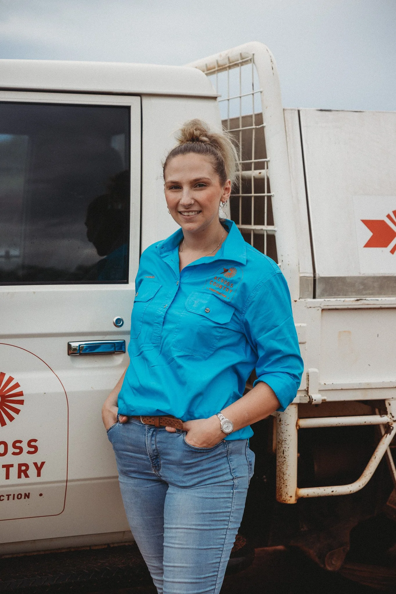A woman standing outdoors next to a white vehicle, wearing a blue shirt with an embroidered logo, with hands in her pockets, smiling at the camera.