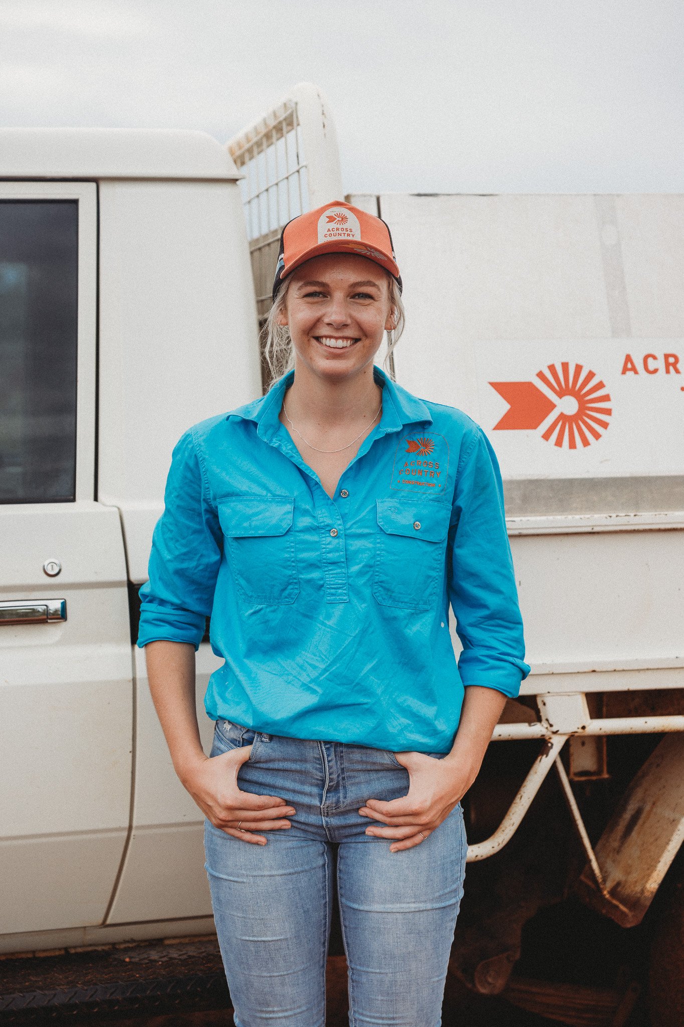 A woman in a blue shirt and jeans standing in front of a white truck with 'Across Country' logo, smiling and hands in her pockets.