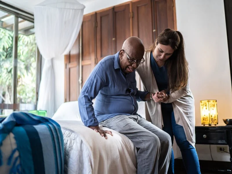 Elderly man sitting on the edge of a bed is carefully assisted by a young female caregiver, who holds his hand and provides support. The home setting emphasizes dignity, safety, and aging in place with wolfmates’ compassionate care.