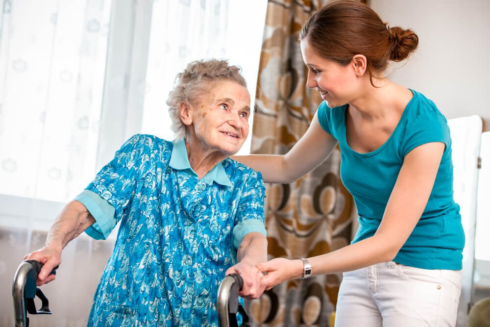 Smiling elderly woman using a walker is assisted by a young female caregiver in a teal shirt, who holds her hand and offers support at home. The scene highlights compassionate in-home care, dignity, and aging in place with wolfmates.