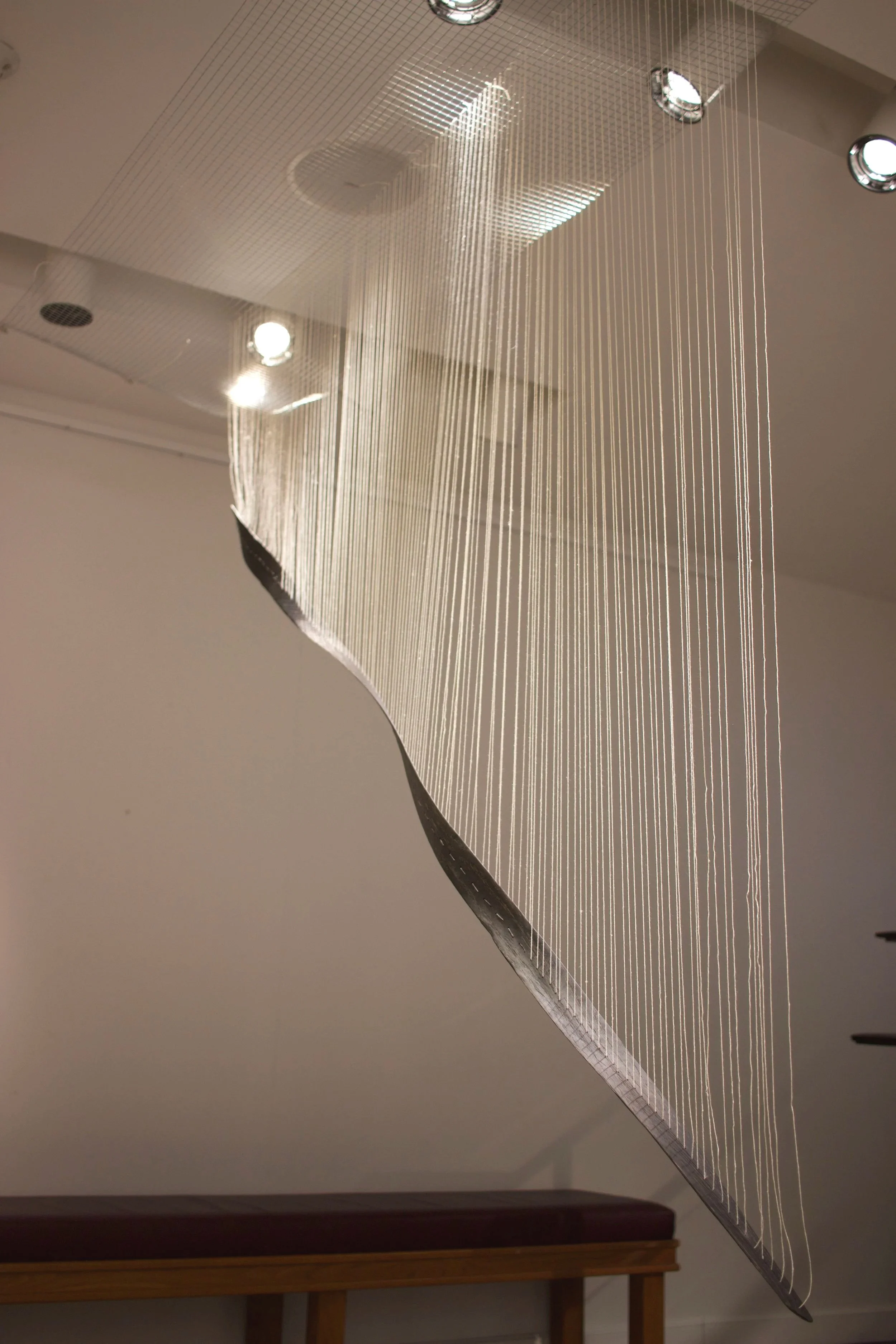 A modern art installation consisting of numerous thin, vertical strings hanging from the ceiling, creating a flowing, wave-like pattern.